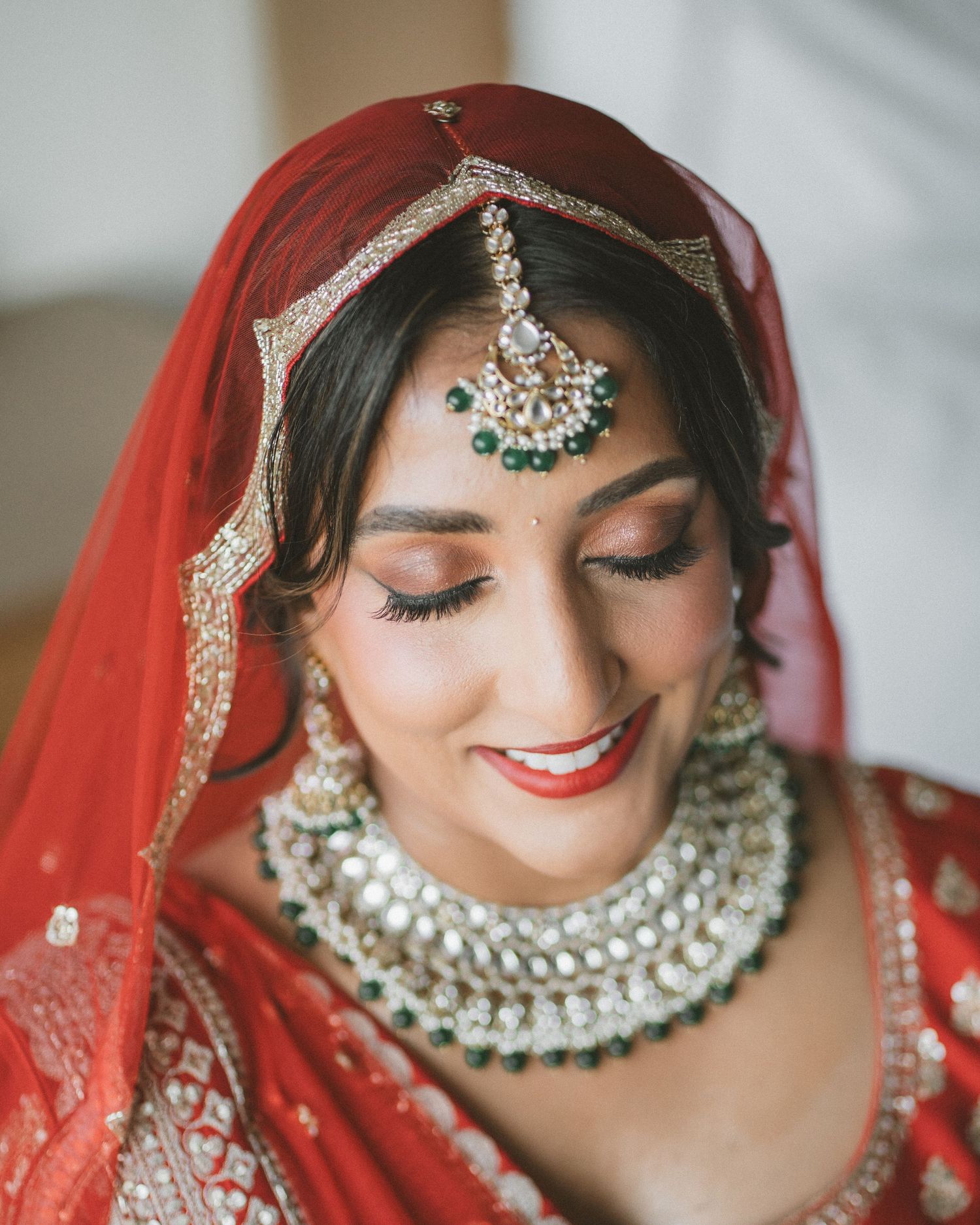 Indian bride in red lehenga with gold maang tikka, emerald jewelry, and pearl necklace smiling gracefully.