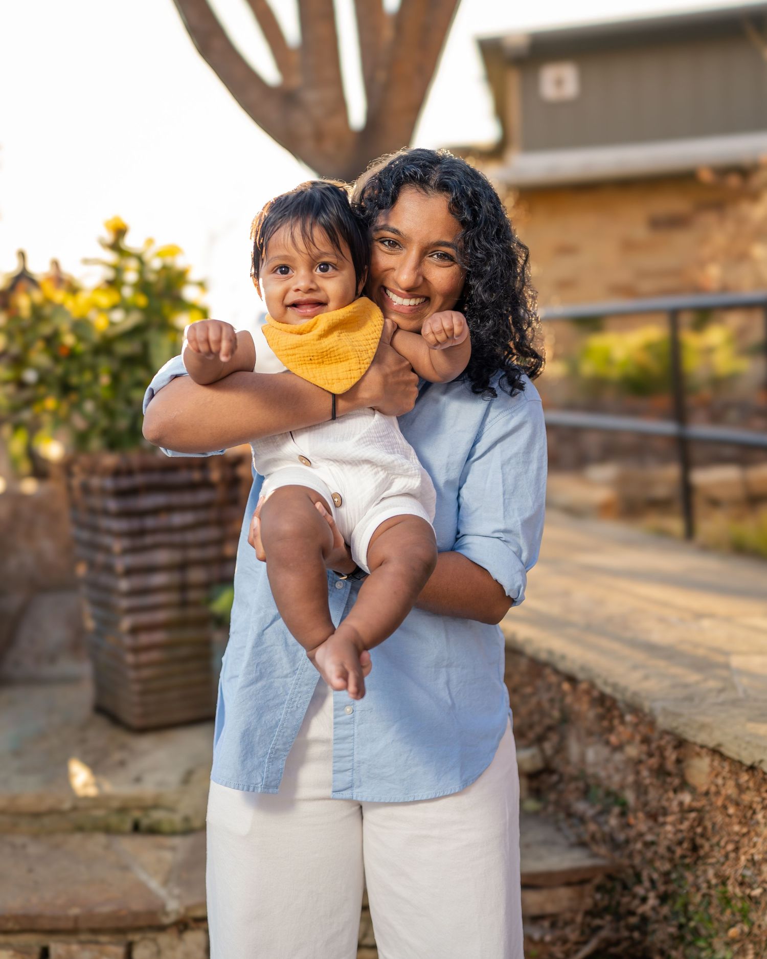 A smiling woman holds a baby wearing a yellow bib outdoors in warm golden light.