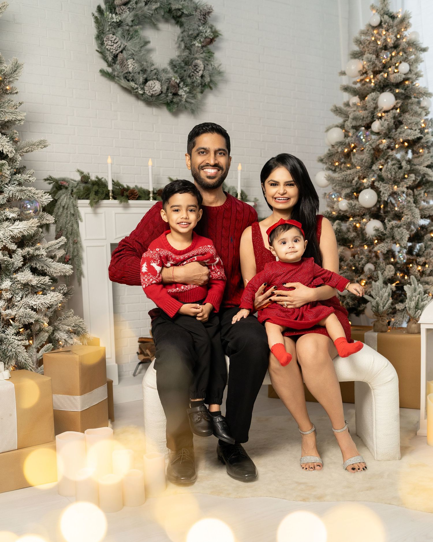 Family of four in matching red outfits posing for a festive Christmas photo with decorated trees and gifts.