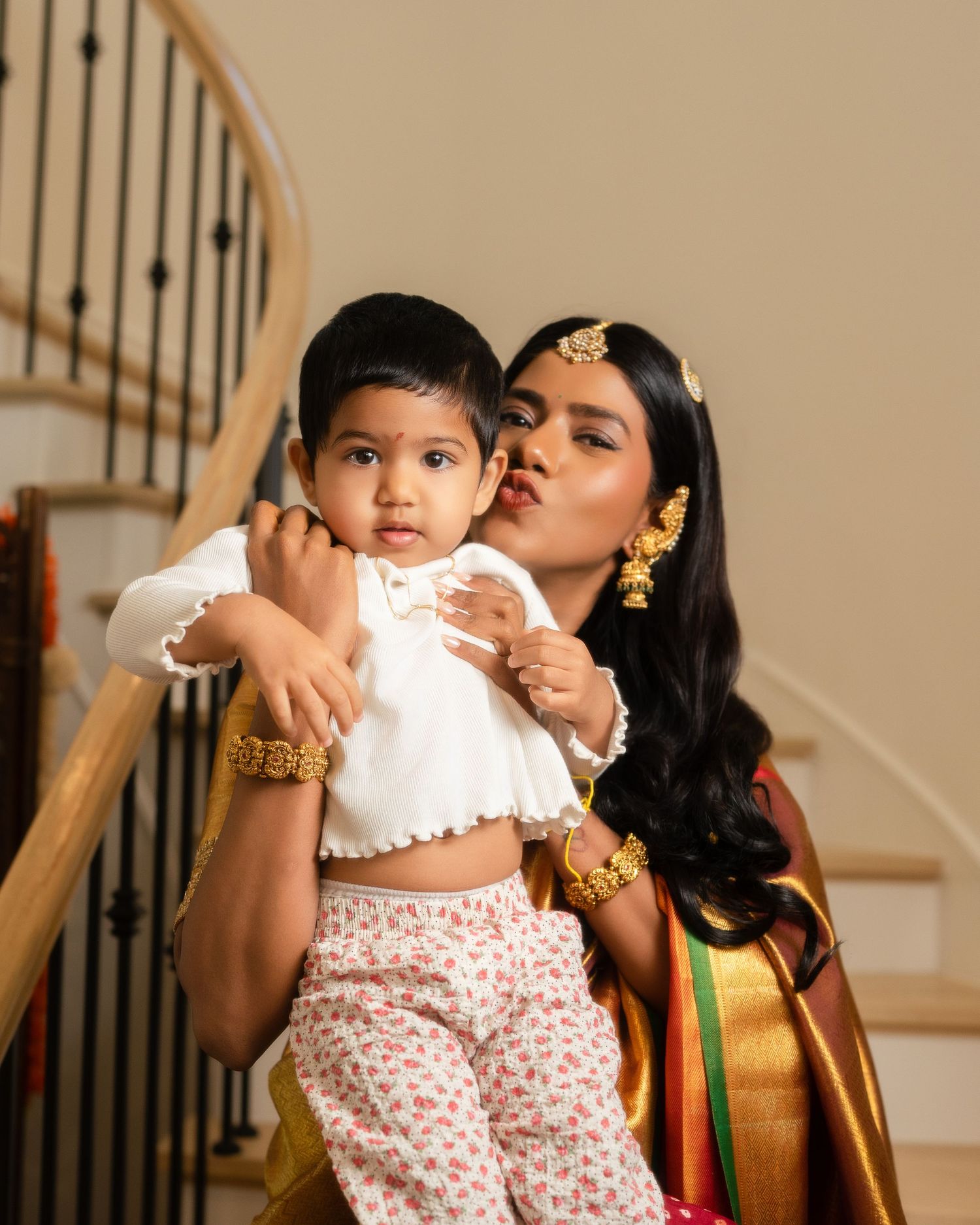 A woman in a green silk saree with gold jewelry kisses a baby in white floral outfit on stairs.