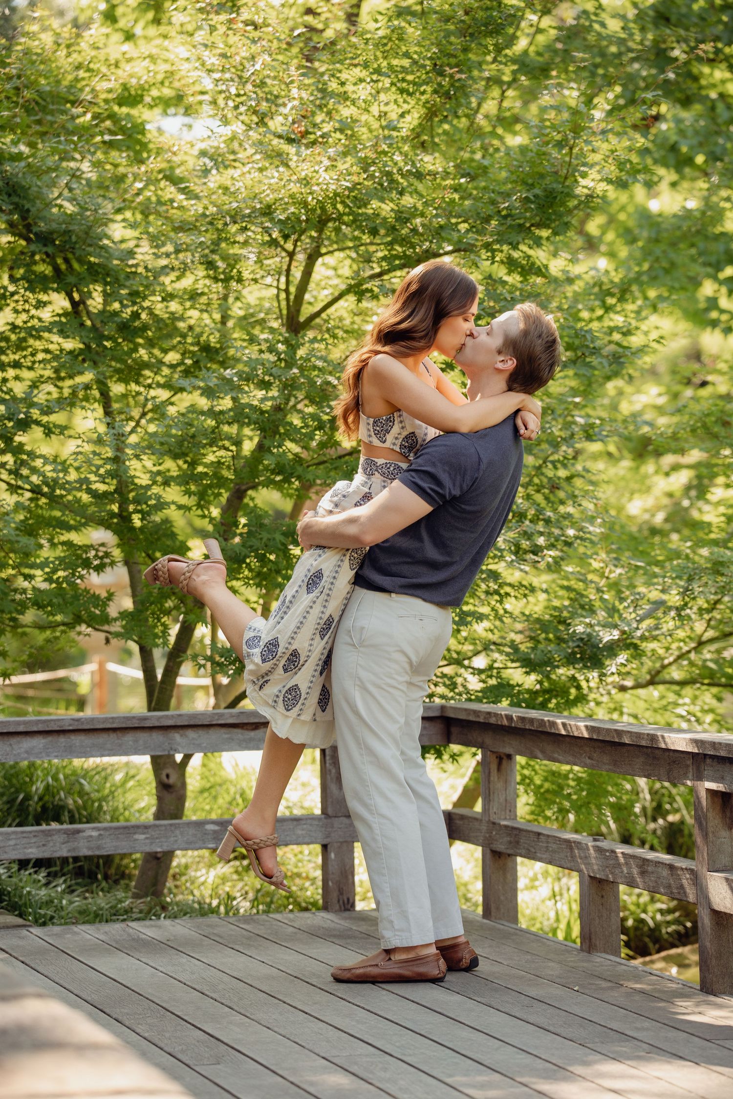 A couple shares a romantic kiss on a wooden bridge surrounded by lush green trees in a garden setting.