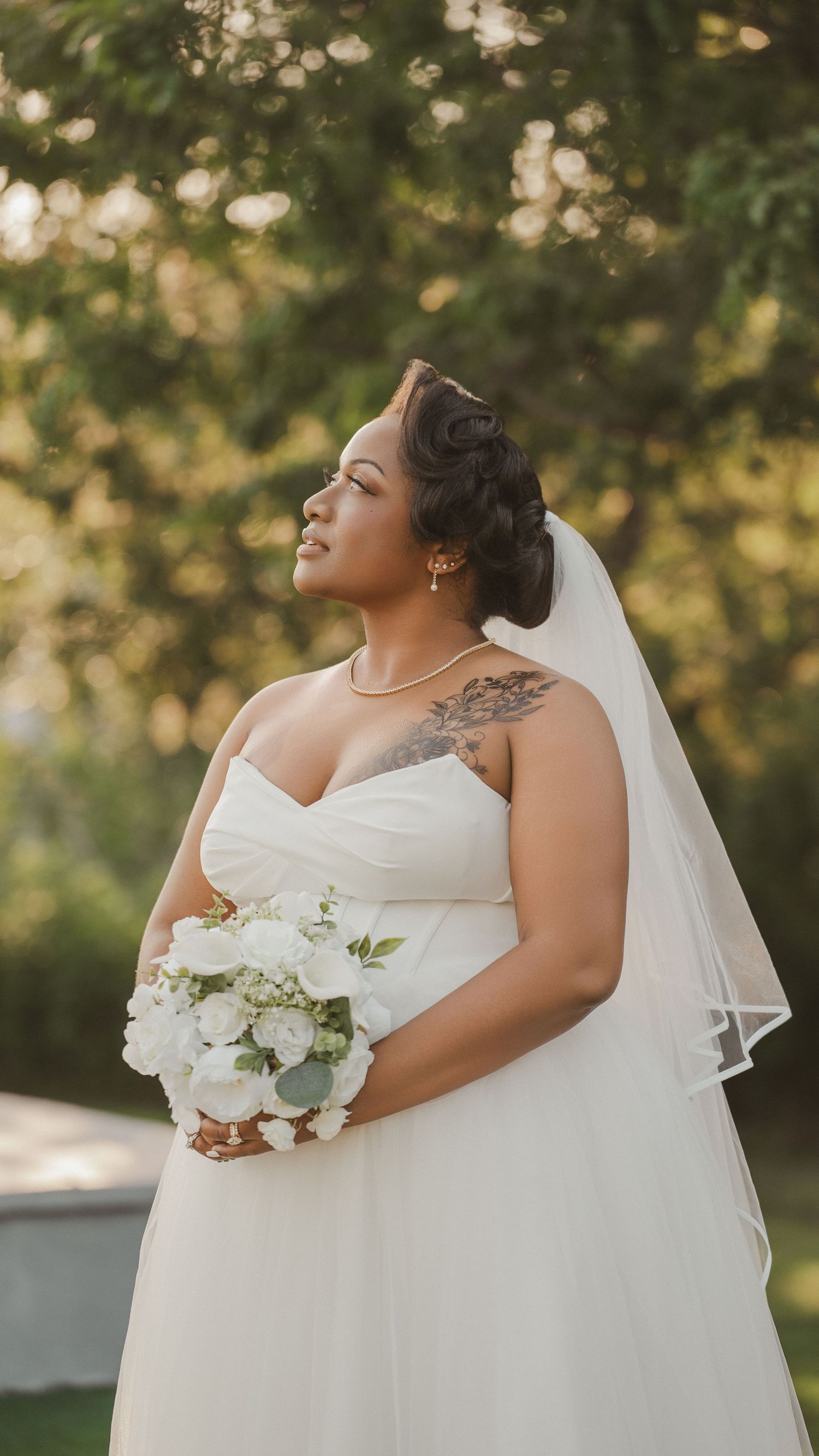 A plus-size bride in a strapless white gown and veil holds a white floral bouquet outdoors among green trees.