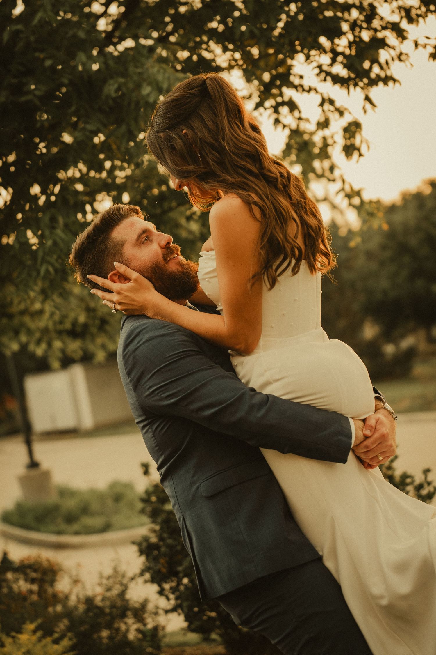 A groom lifts his bride in a white dress during a romantic golden hour outdoor wedding photo.
