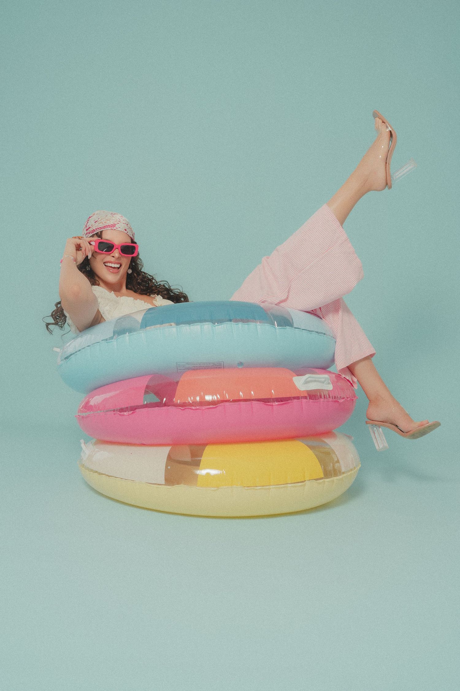 Happy woman in pink outfit sitting in stacked colorful inflatable pool rings on light blue background.