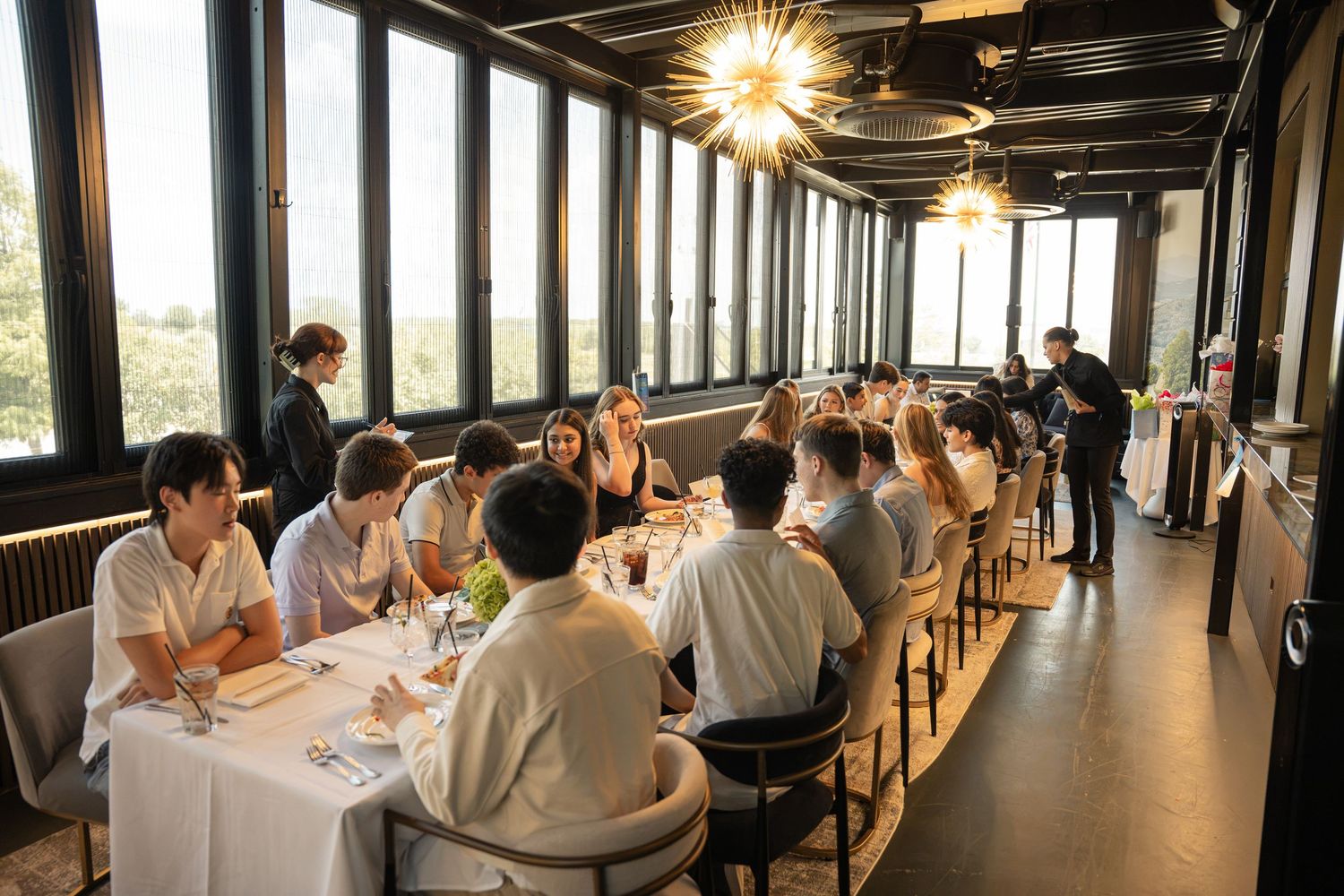 Elegant private dining room with long table, guests enjoying a meal, large windows, and modern gold chandelier lighting.