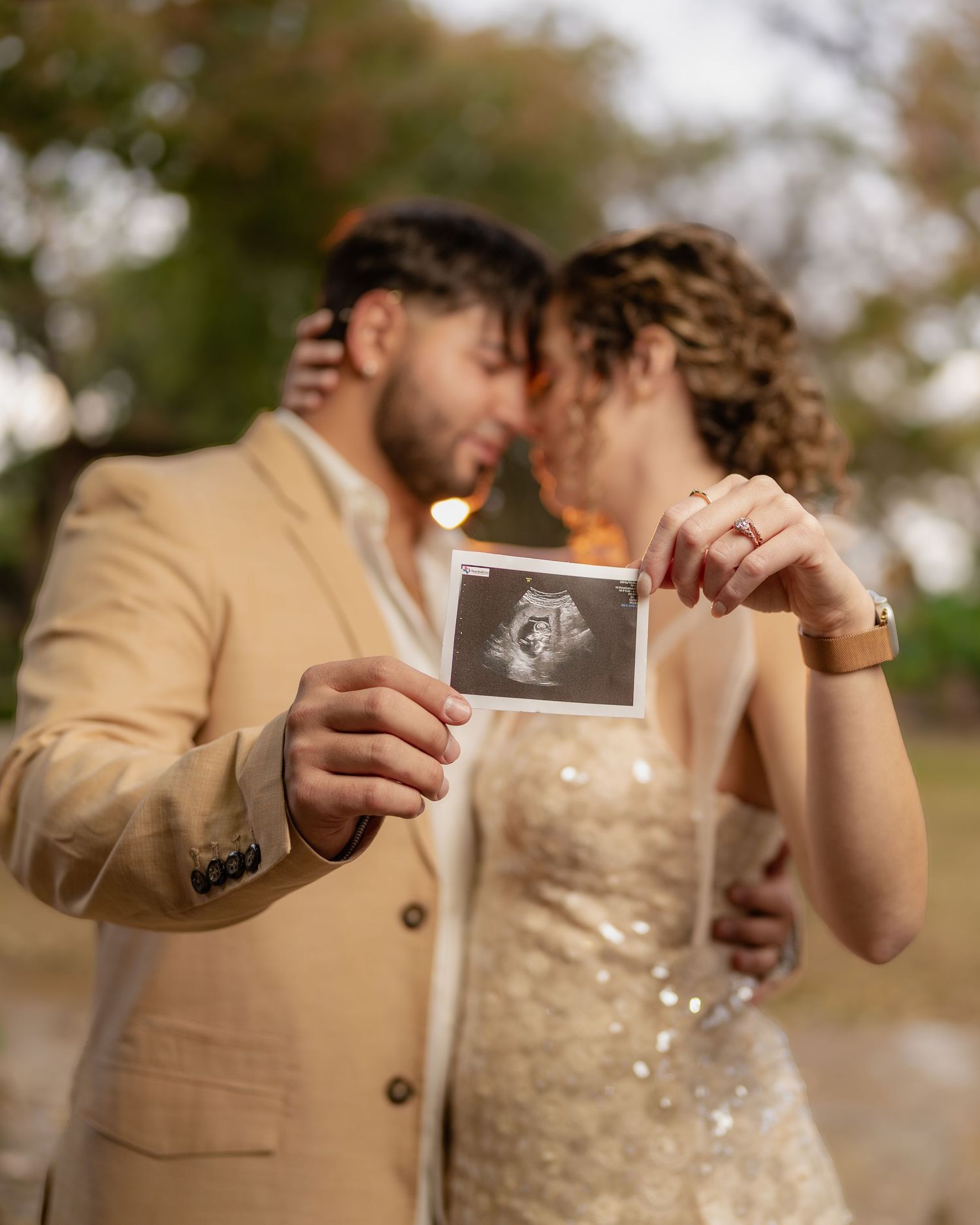 Couple in beige and gold outfits sharing emotional moment while holding ultrasound photos.