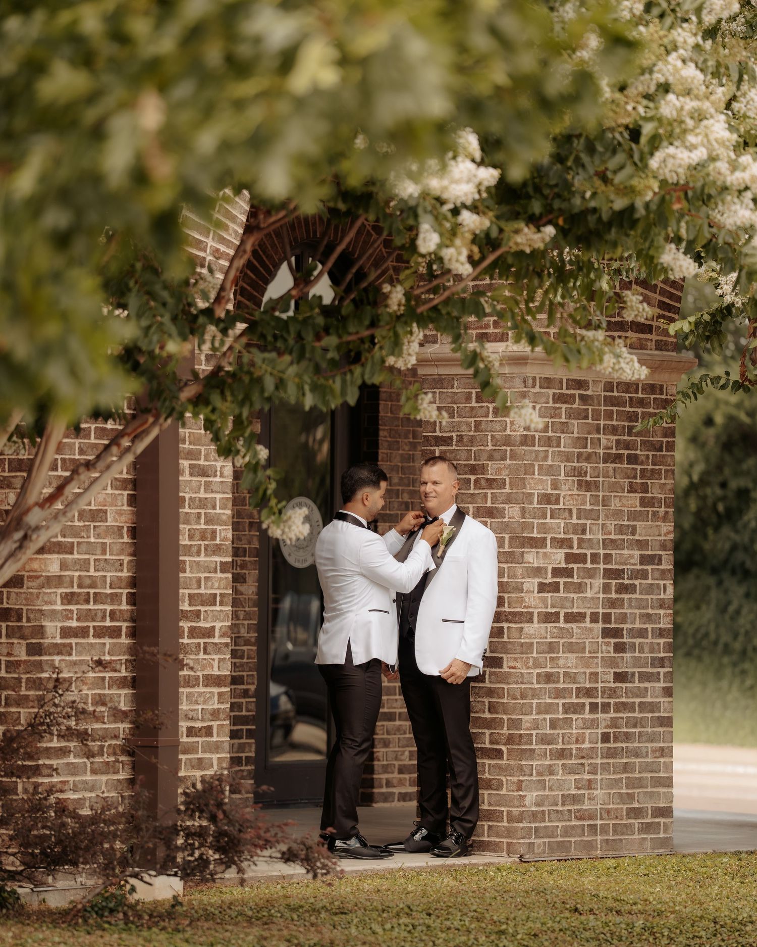Two grooms in white tuxedo jackets share a tender moment outside a brick building surrounded by blooming trees.