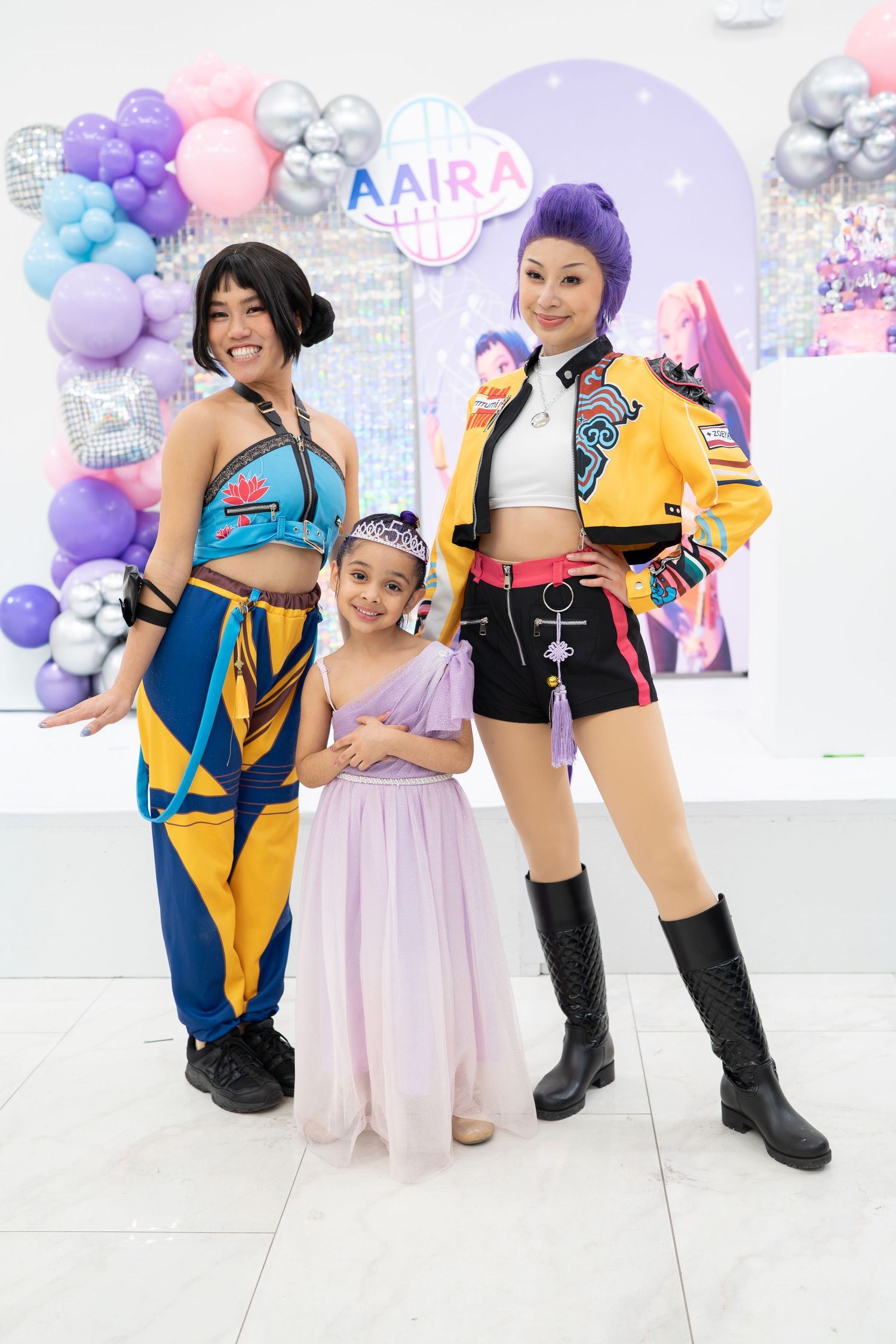 Cosplayers in vibrant yellow and blue outfits pose playfully against a pastel balloon backdrop at a convention.