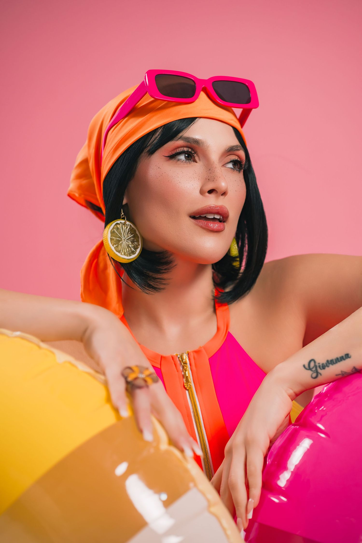 Woman in pink swimsuit, orange headscarf, pink sunglasses, and lemon earrings holding colorful inflatable balls.