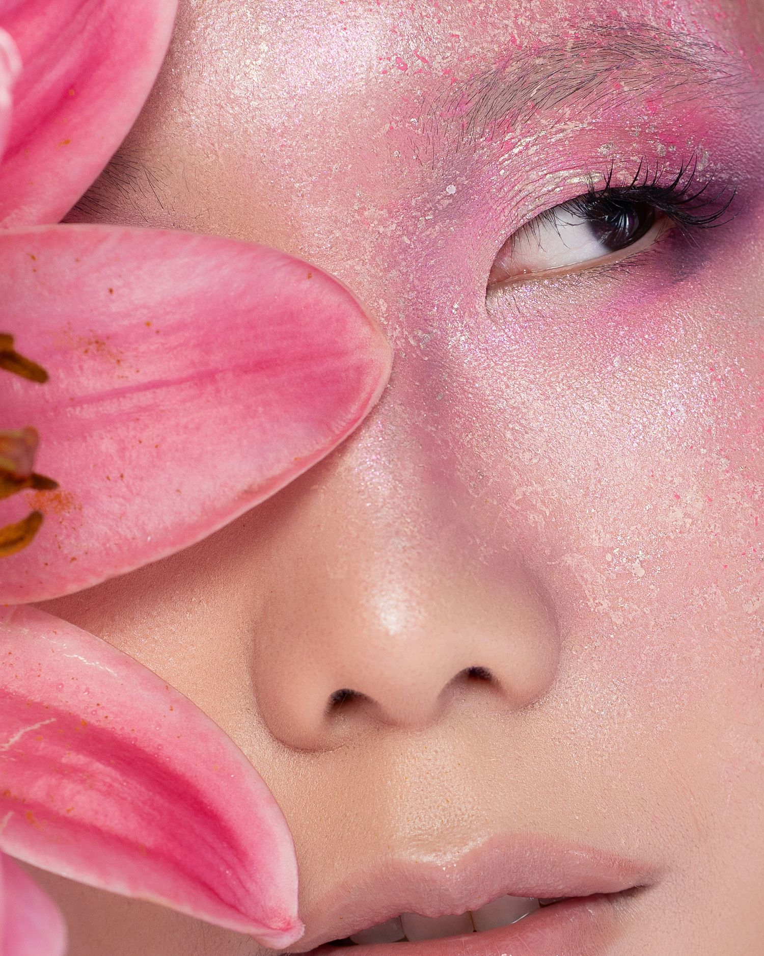 Close-up of a model wearing pink glittery eye makeup with a pink lily flower beside her face.