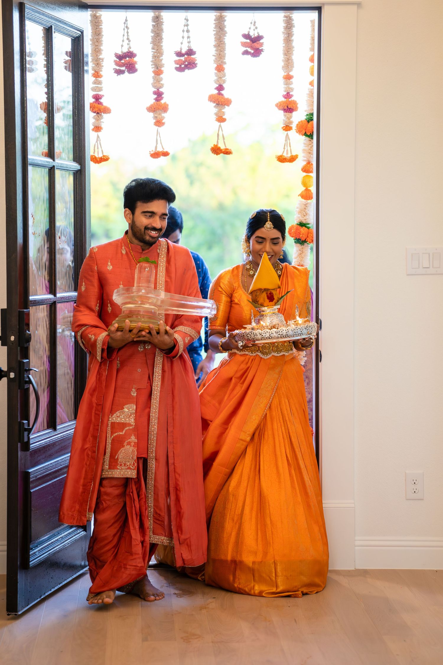 Indian couple in traditional orange attire carrying ritual items during a Hindu wedding ceremony with floral decorations.