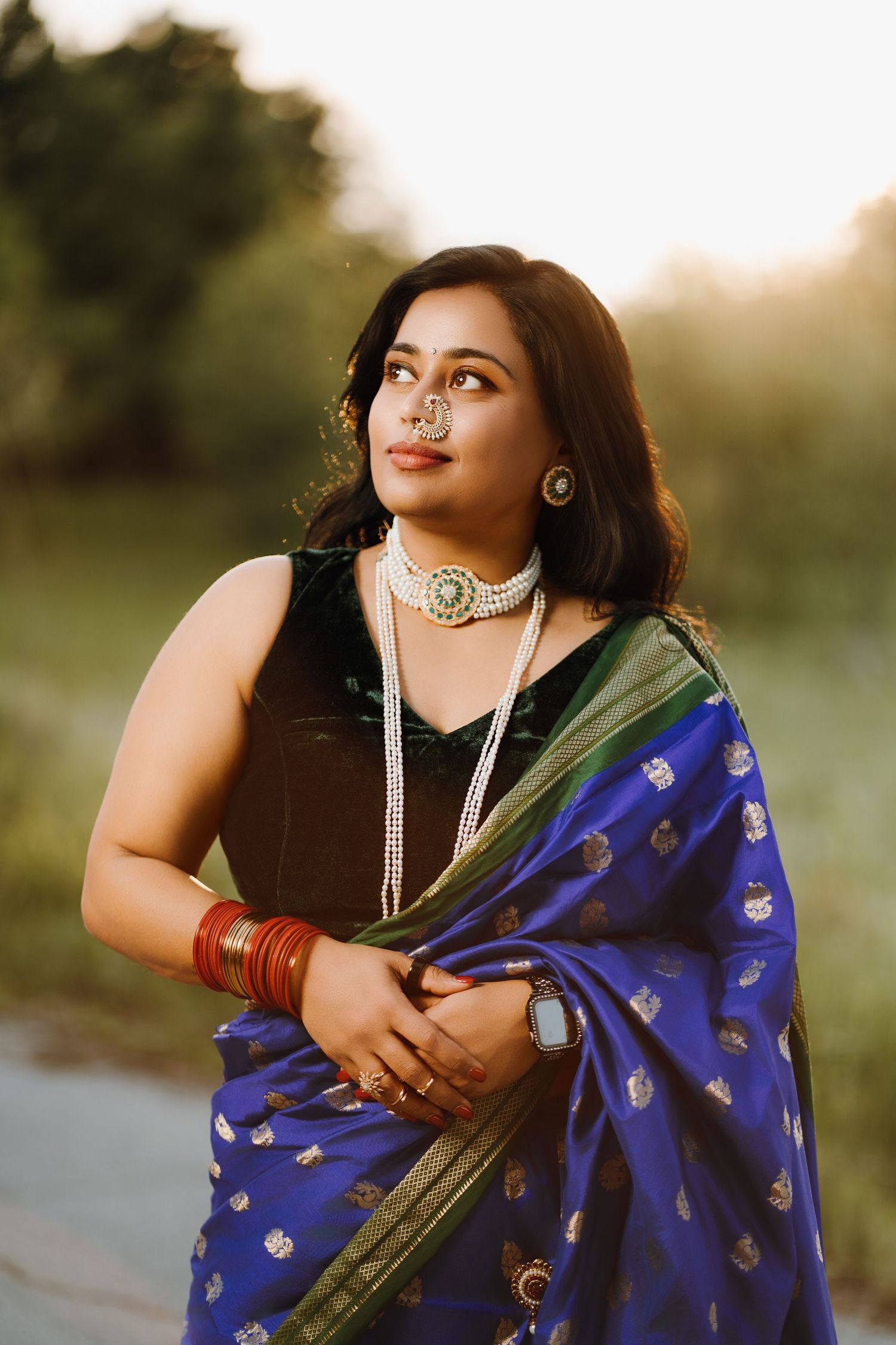Woman in blue silk saree with gold motifs, pearl choker, nose ring, and red bangles poses outdoors at golden hour.