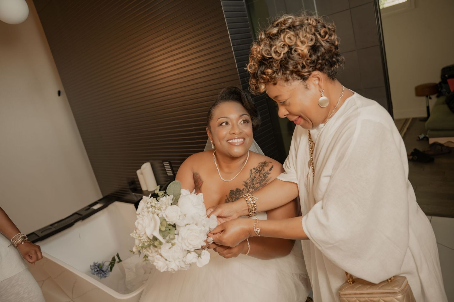 Bride in white gown holding bouquet smiles as woman helps her with bracelet before wedding ceremony.