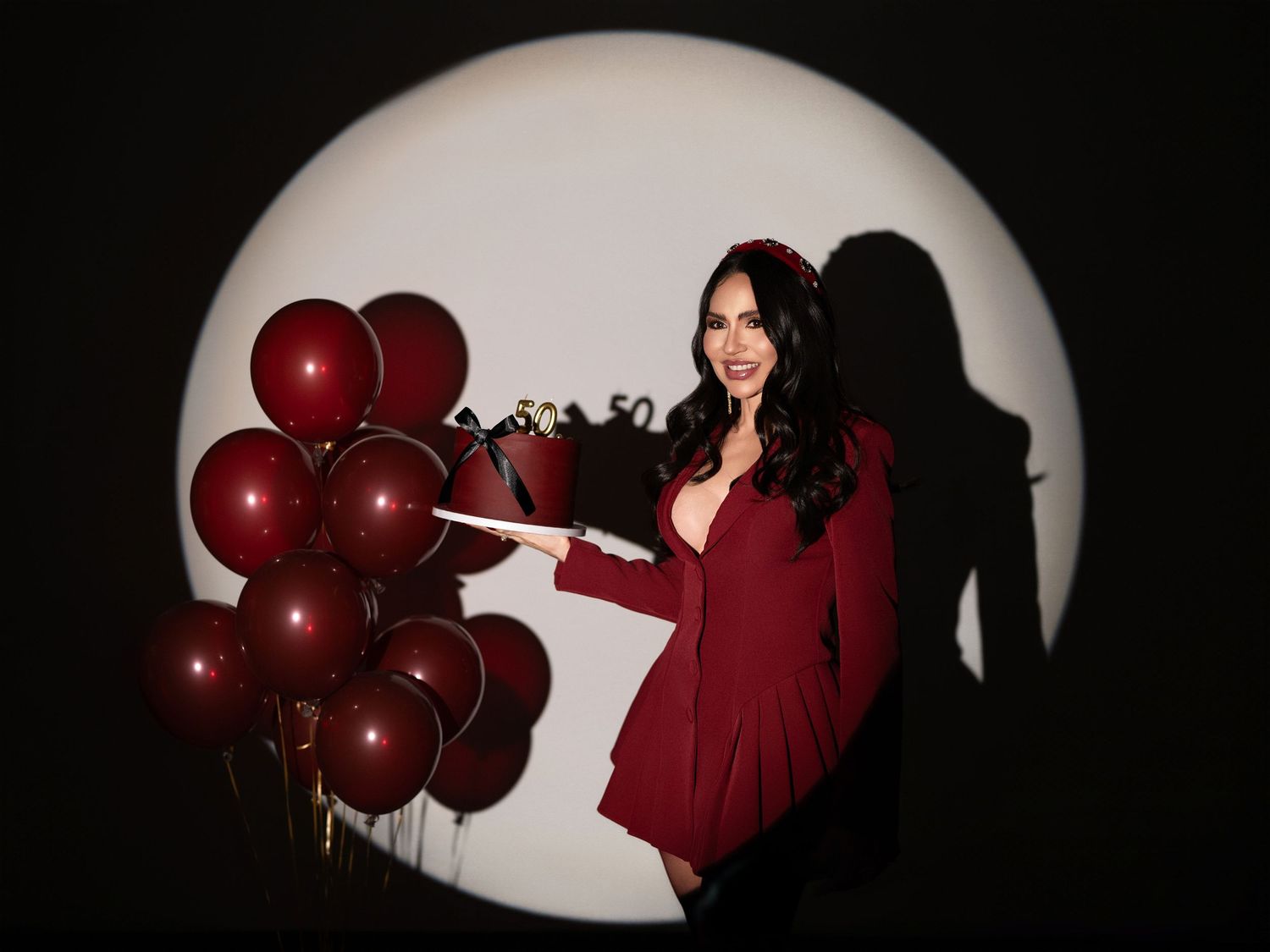 Woman in red dress holding dark red balloons poses in front of glowing white circle backdrop.
