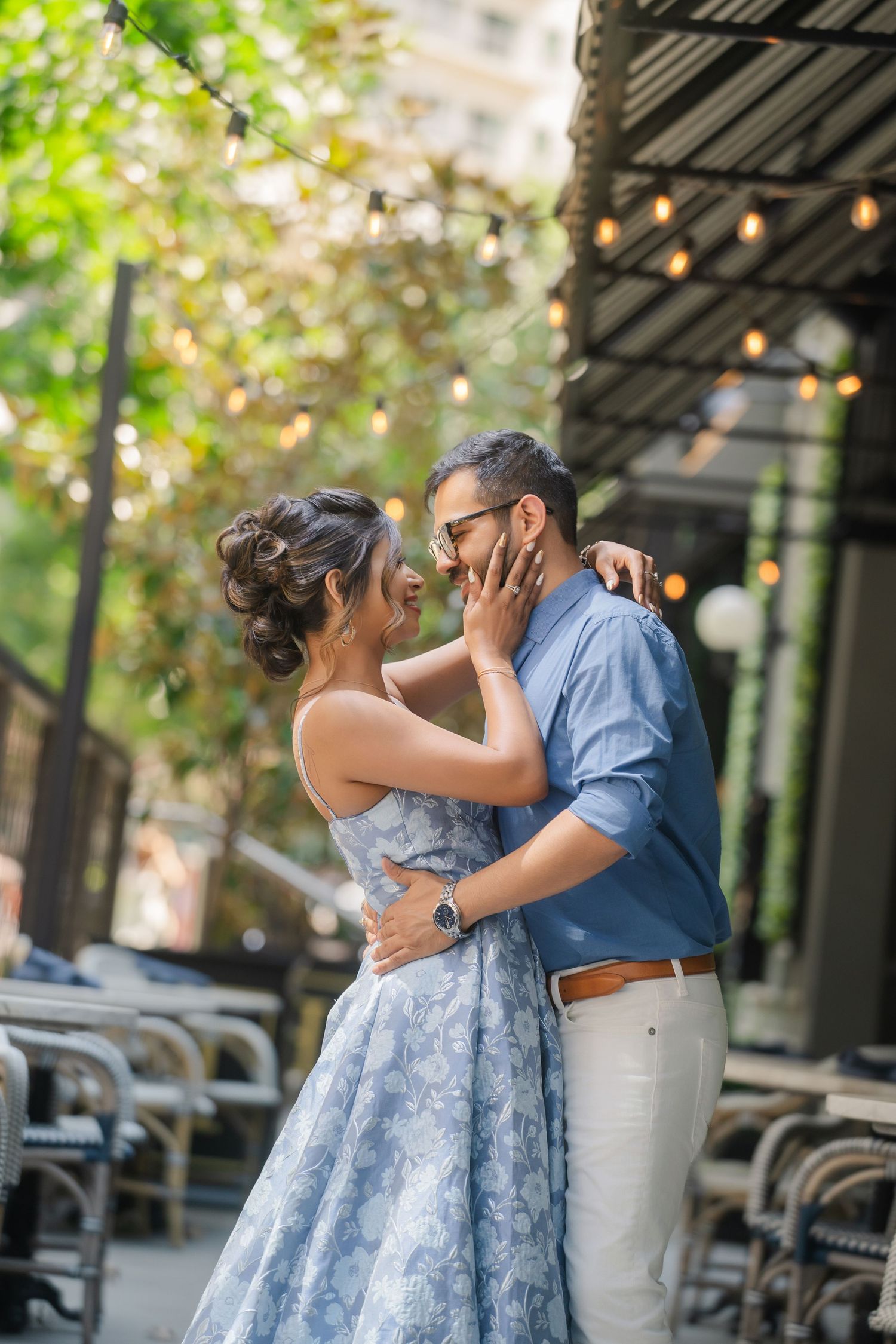 Couple sharing a romantic moment outdoors, woman in blue floral dress touching man's face, string lights in background.