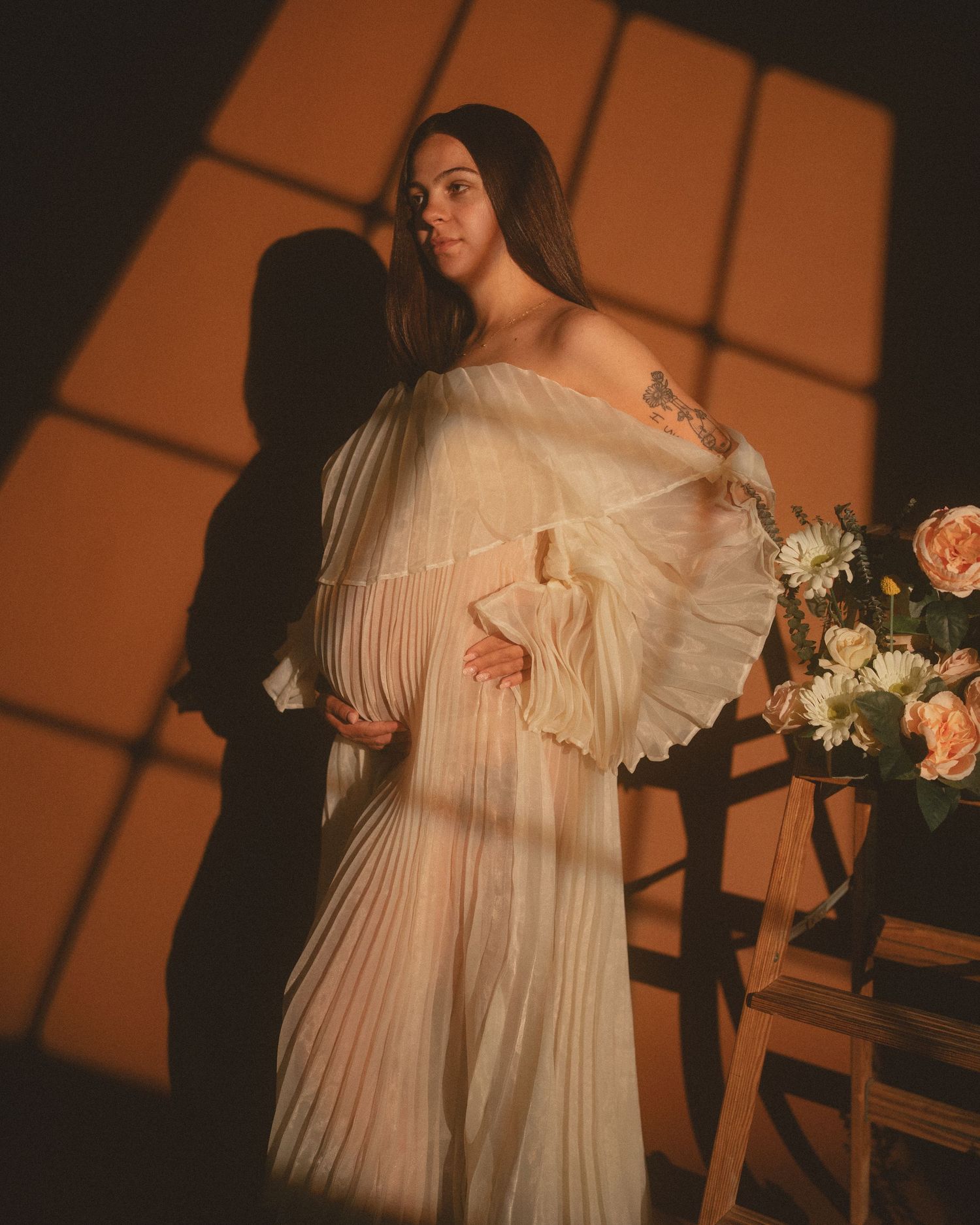 Pregnant woman in white off-shoulder pleated gown poses elegantly beside flowers in warm golden light.
