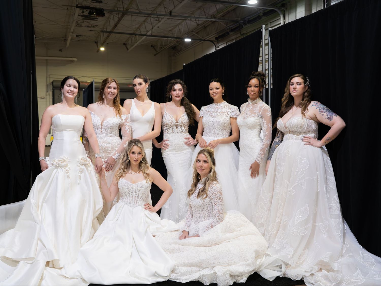 Group of women modeling elegant white bridal gowns at a wedding expo, posing together in a dark studio setting.