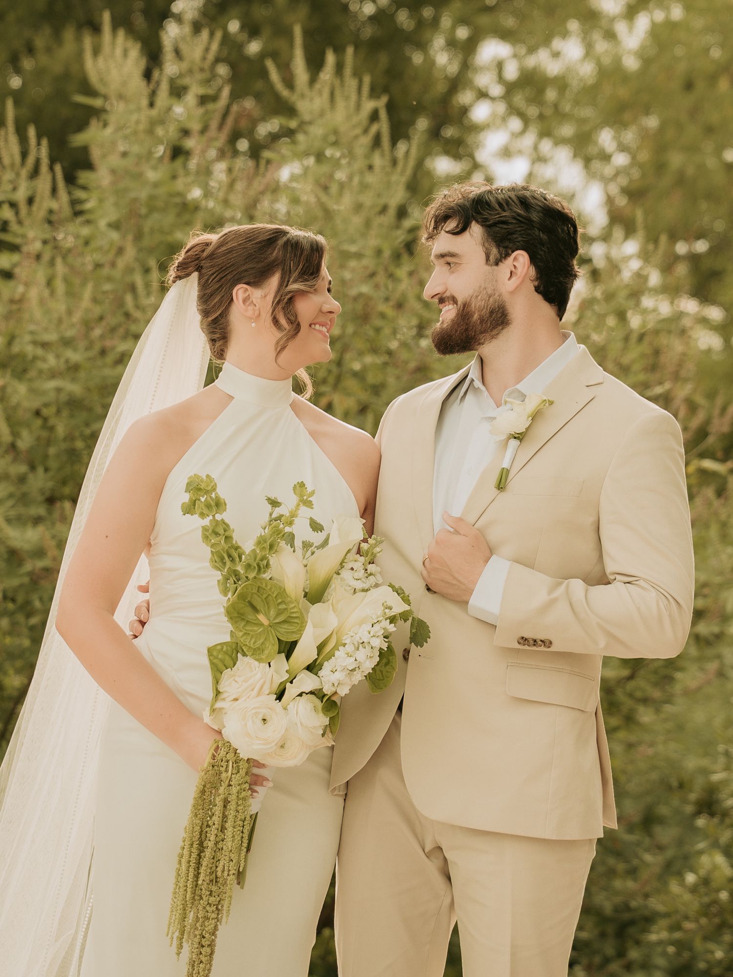 A bride and groom share a loving glance outdoors, she holds a white bouquet, he wears a tan suit.