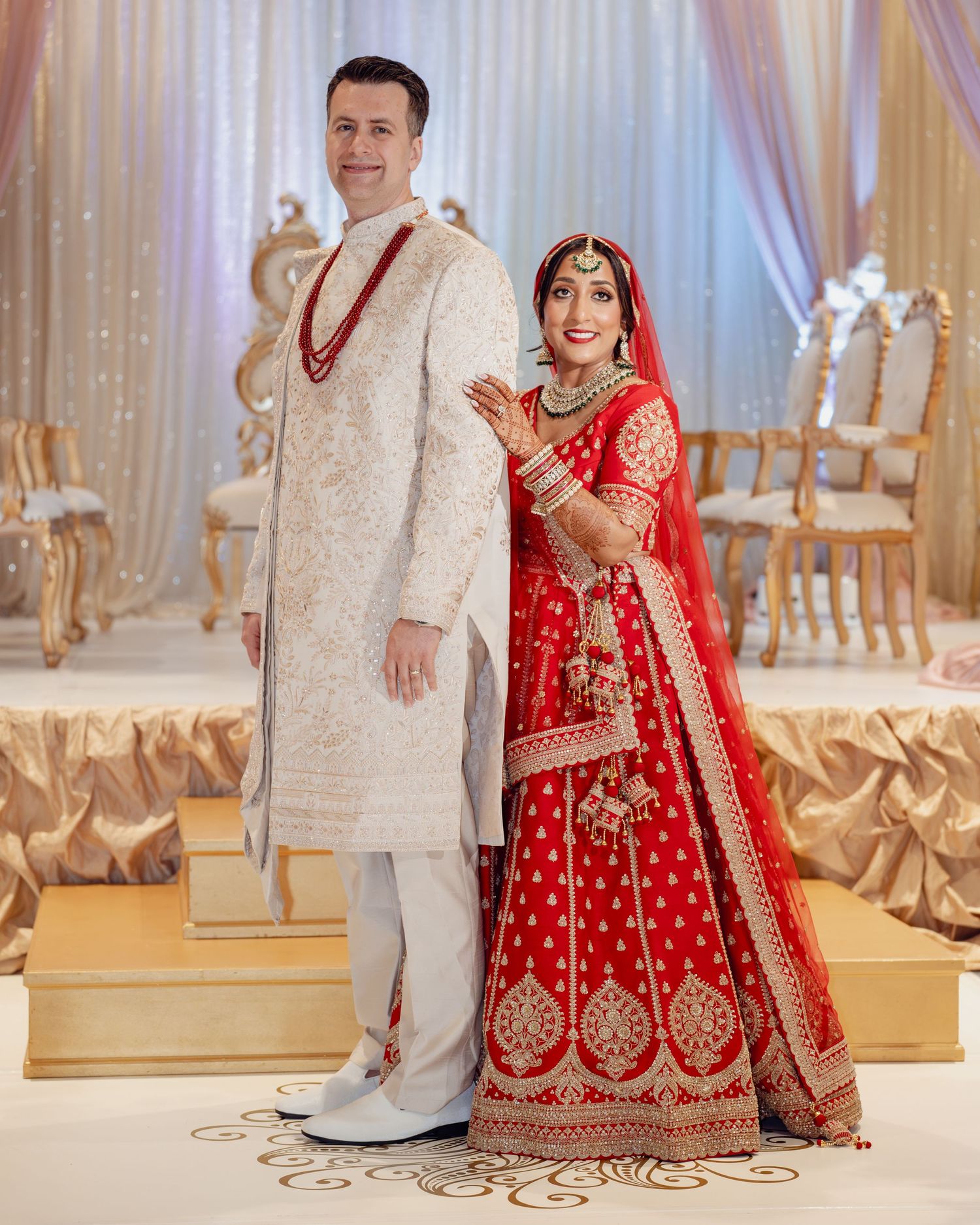 Couple in traditional Indian wedding attire, groom in white sherwani, bride in red lehenga with gold embroidery.