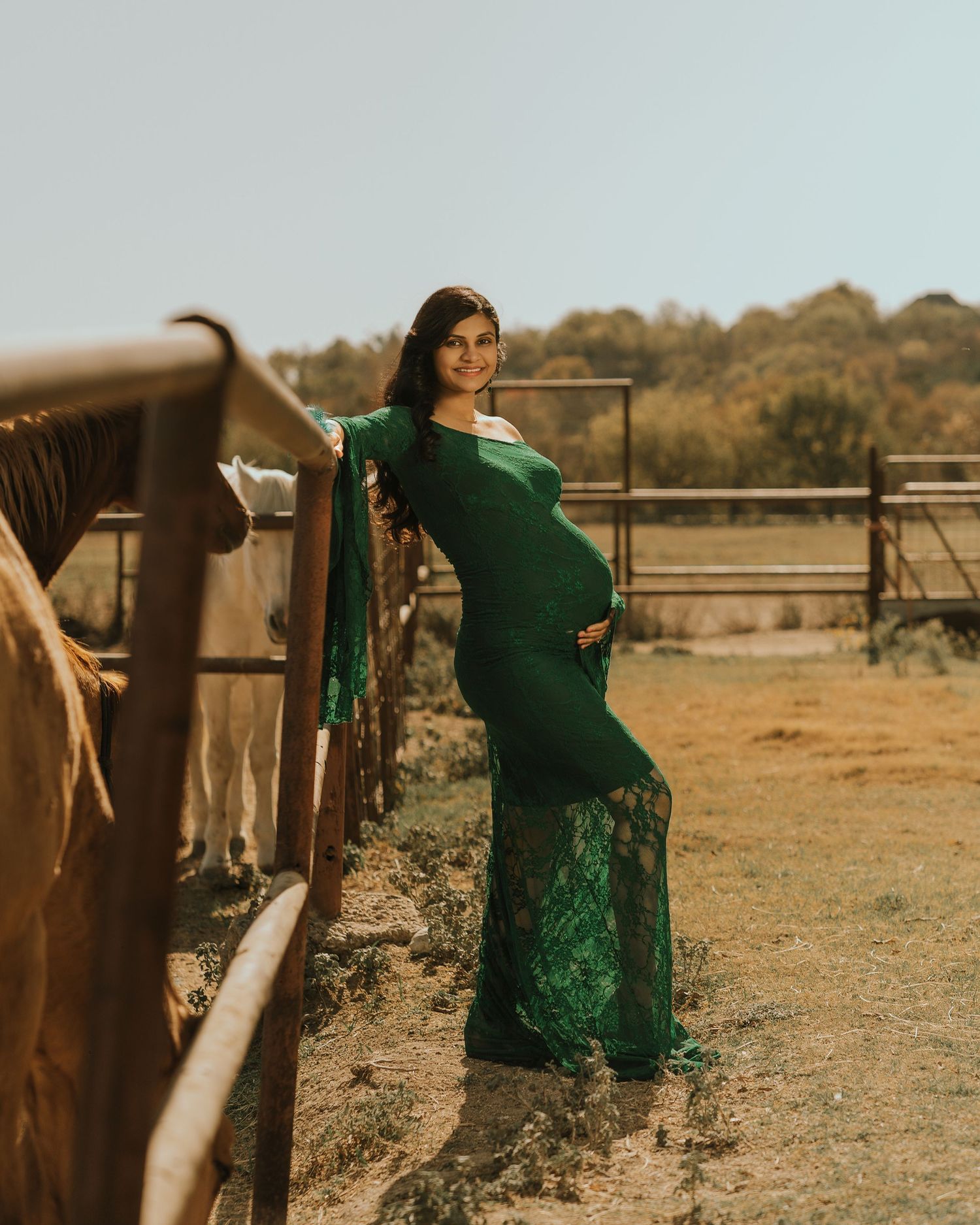 Pregnant woman in elegant green lace gown posing by a rustic fence at a farm during a maternity photoshoot.
