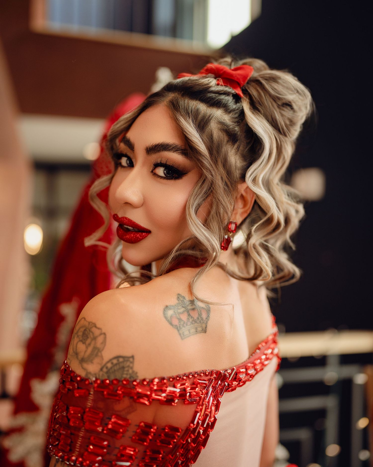 Woman in red sequin off-shoulder dress with crown tattoo, red veil, bold red lips, and curly updo hairstyle.