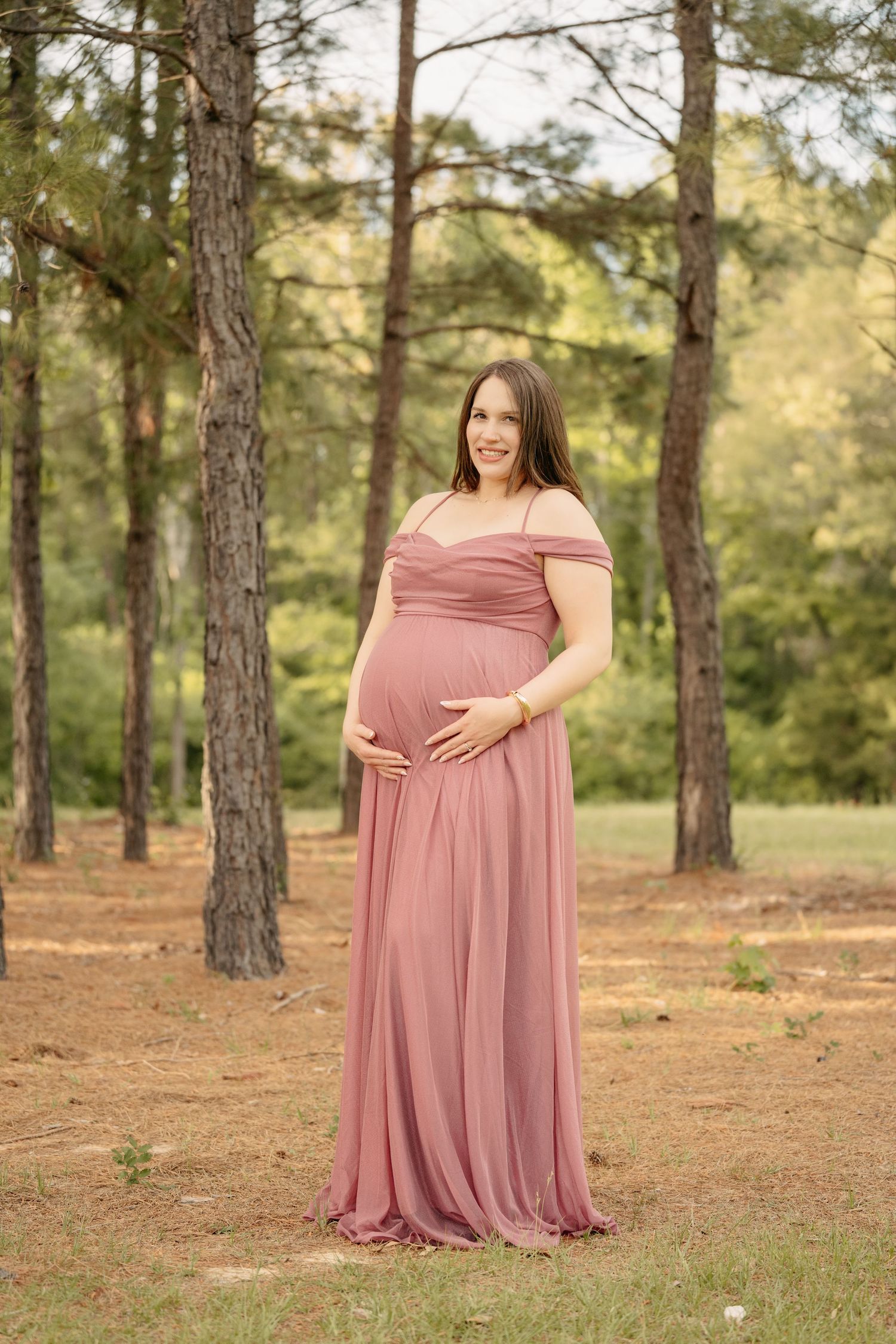 Pregnant woman in pink maxi dress holding belly outdoors in pine forest for maternity photo session.