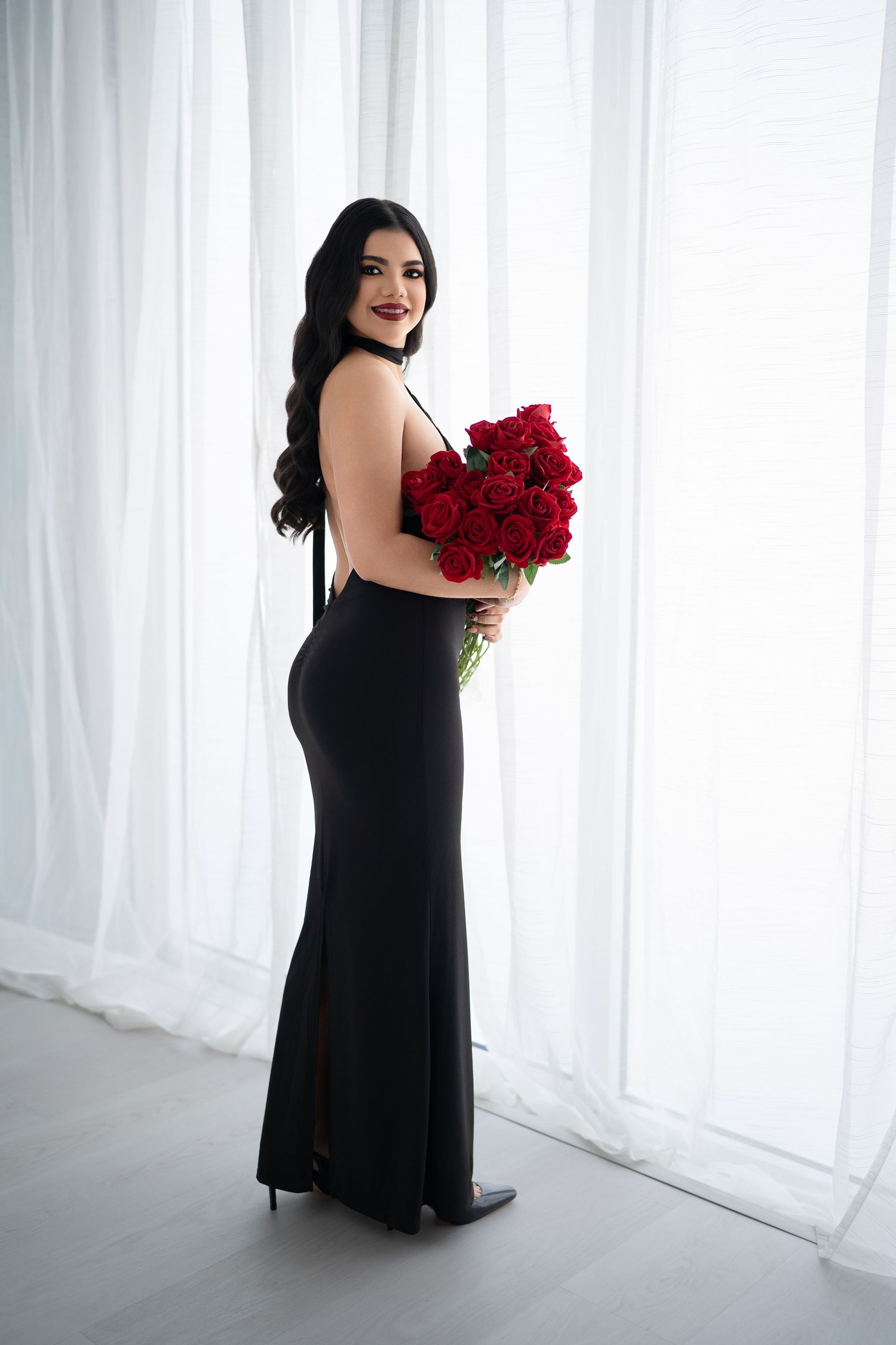 Elegant portrait session with black dress and red roses against white curtain backdrop.