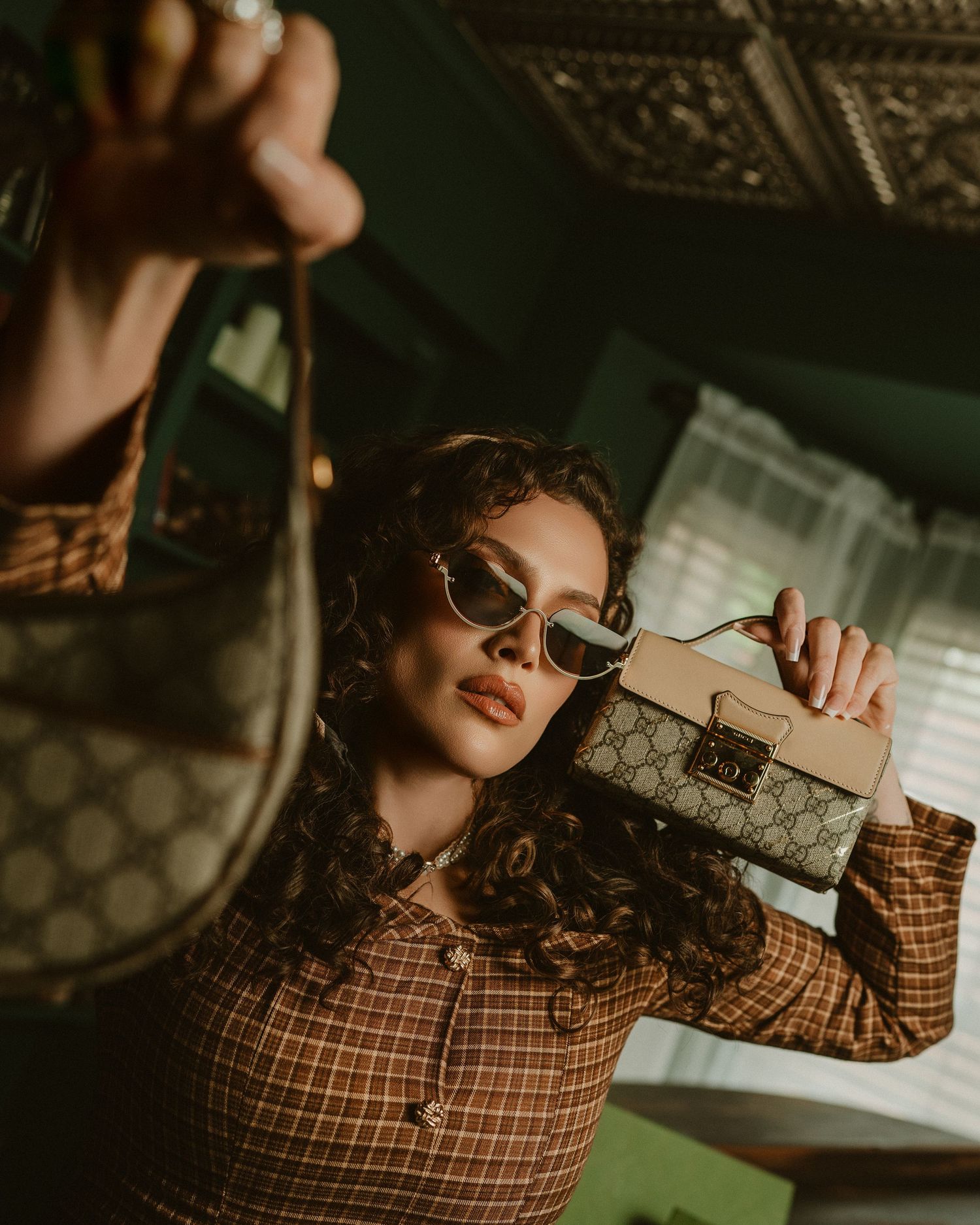 Woman in retro plaid shirt and sunglasses holding two Gucci handbags in a vintage-styled room.