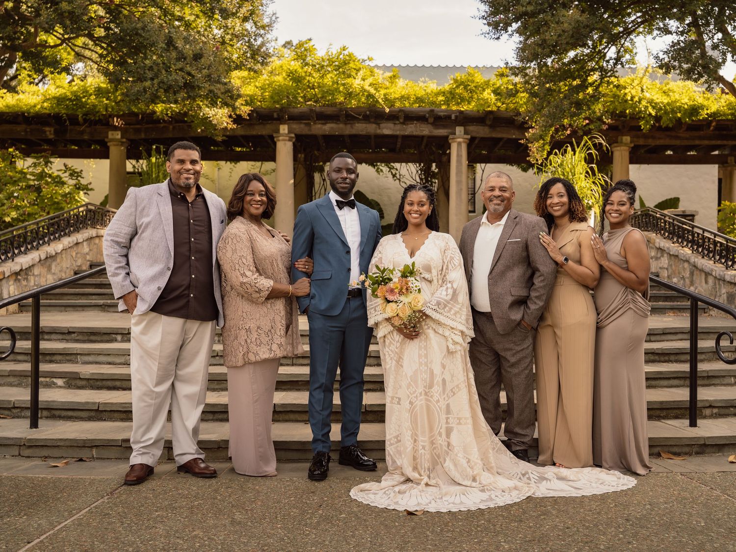 Wedding party poses on stone steps, bride in lace gown with bouquet, groom in blue suit, flanked by family.