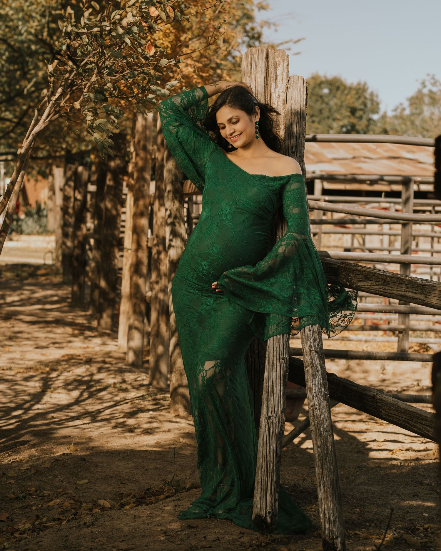 Woman in elegant green lace gown posing by rustic wooden fence in outdoor farm setting.