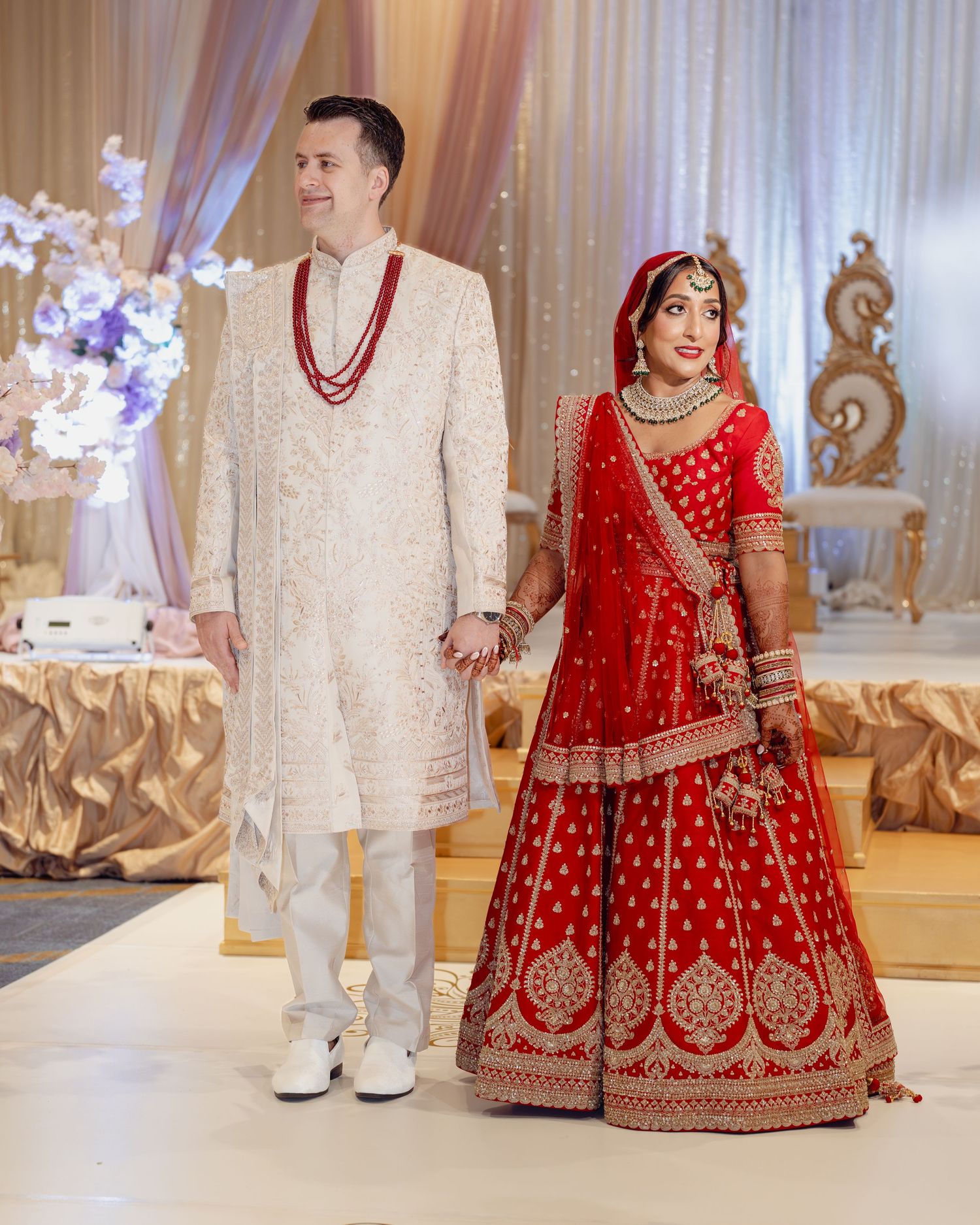 Bride in red lehenga and groom in white sherwani hold hands at elegant Indian wedding ceremony.