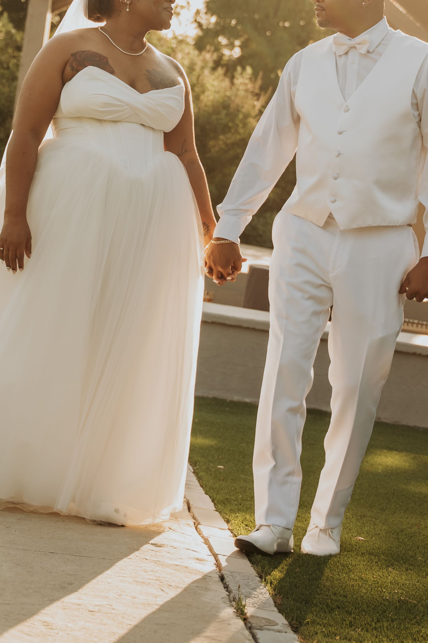 Bride and groom holding hands in white wedding attire, walking on grass in golden sunset light.