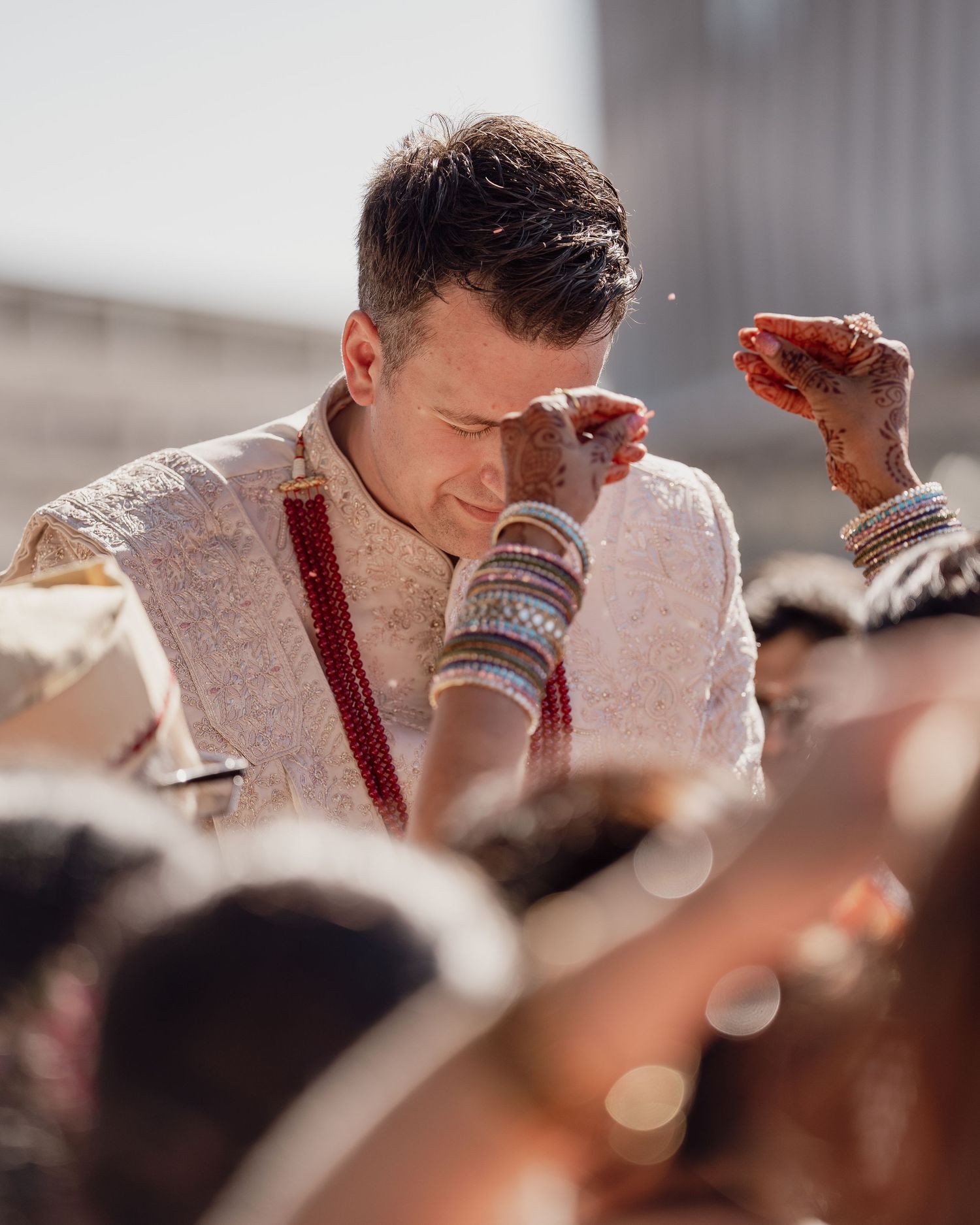 Groom in white sherwani with red dupatta and bangles bowing during Indian wedding ceremony outdoors.