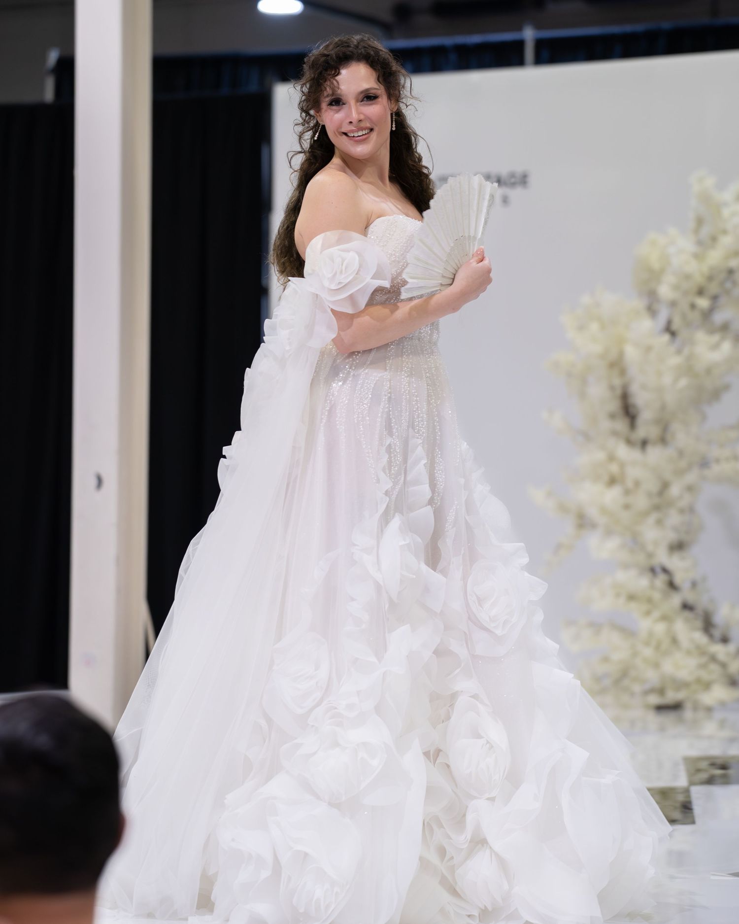A model in an off-shoulder white floral ball gown holding a fan at a bridal fashion show.