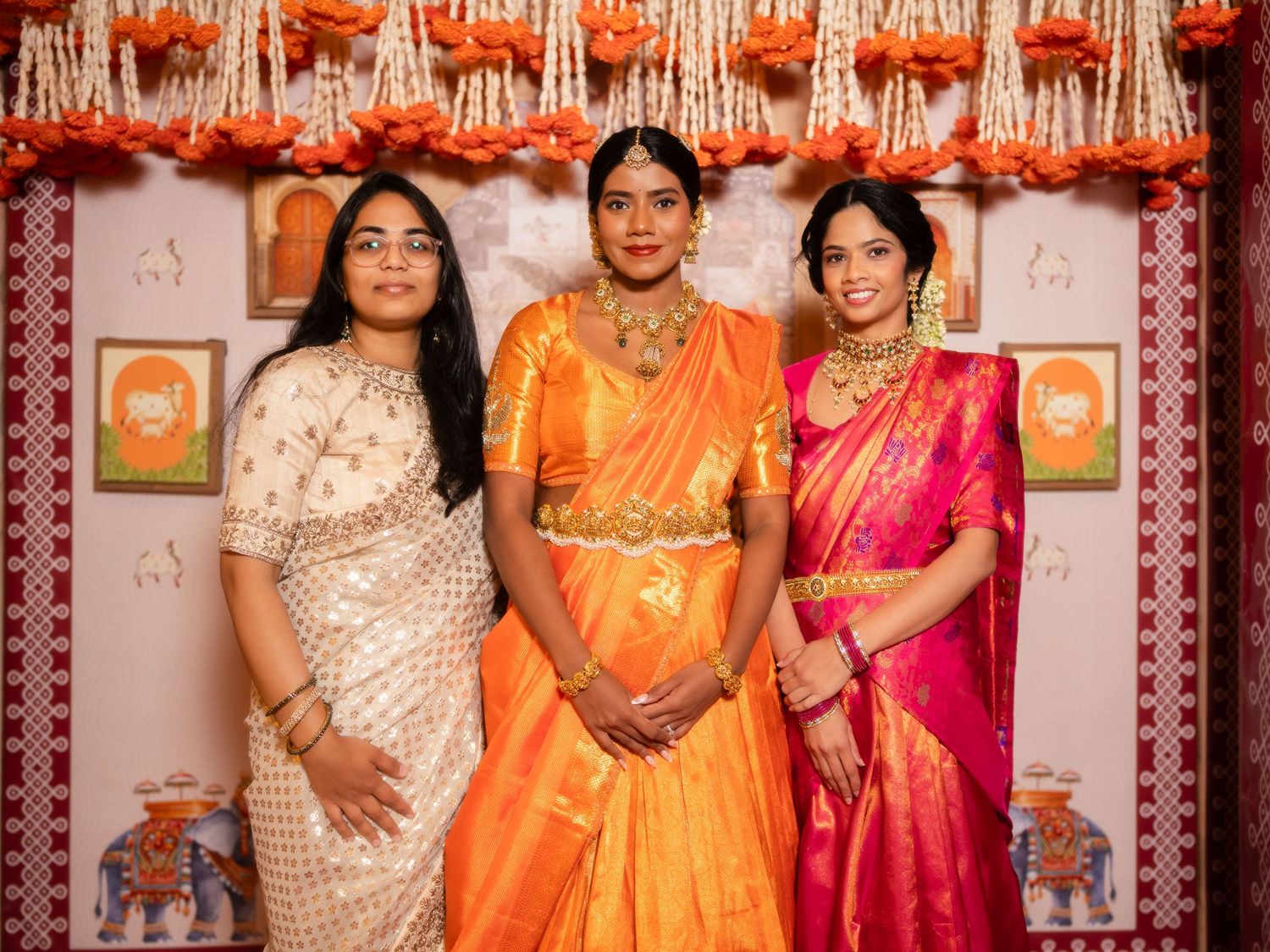 Three women in vibrant traditional Indian sarees pose together at a festive celebration decorated with marigold garlands.