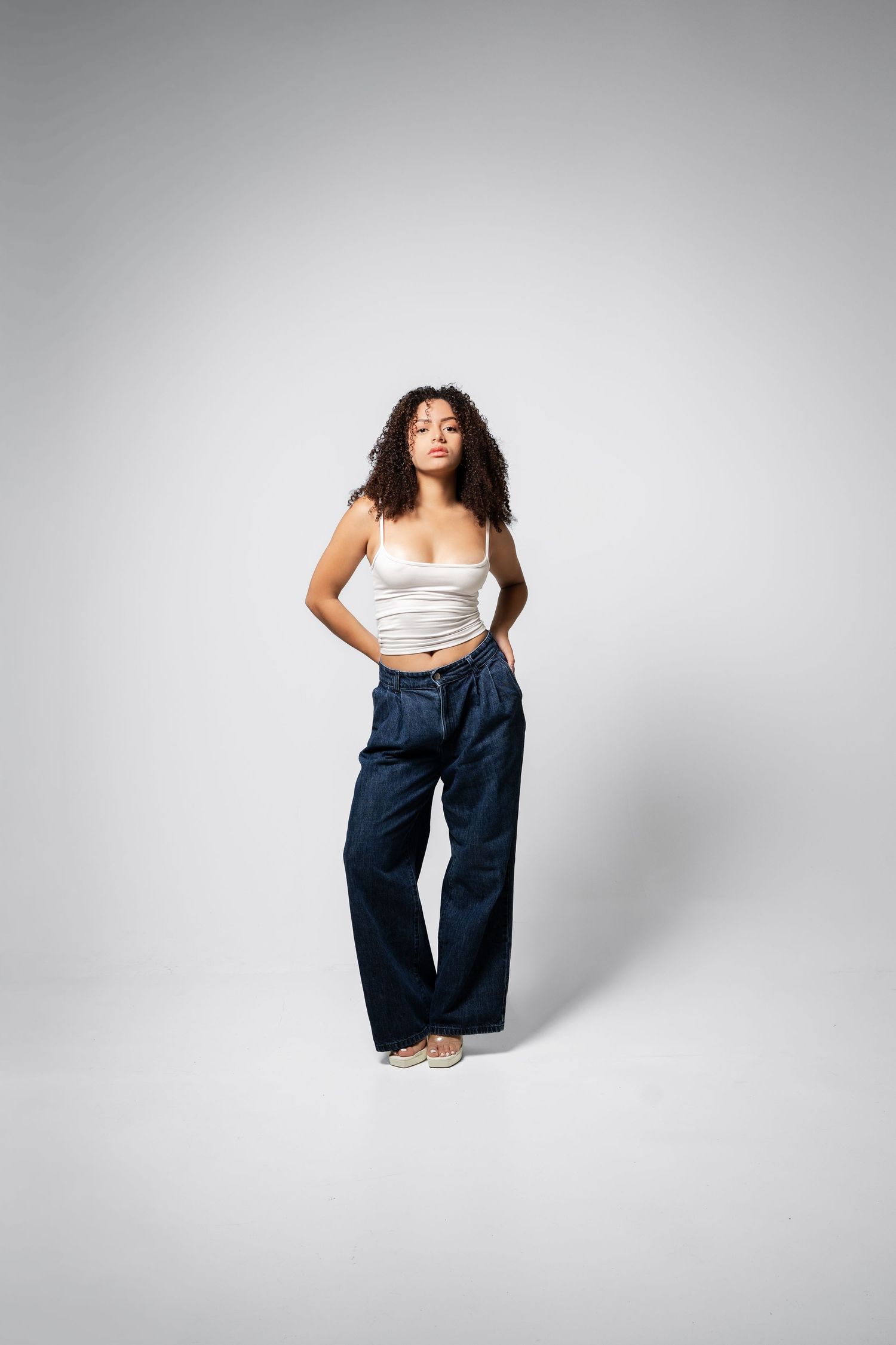 Woman in white crop top and wide-leg dark jeans posing against a light gray background.