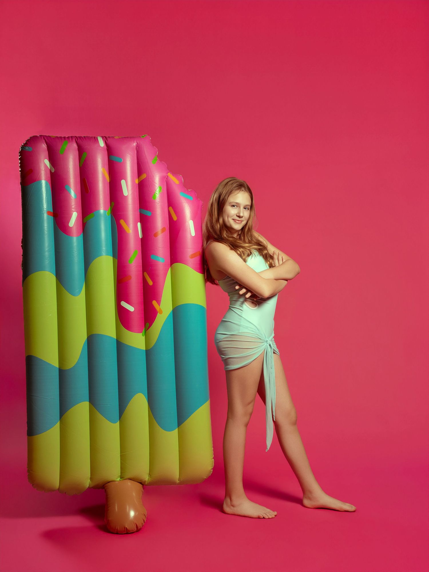 Young woman in mint swimsuit poses with colorful donut inflatable float against vibrant pink background.