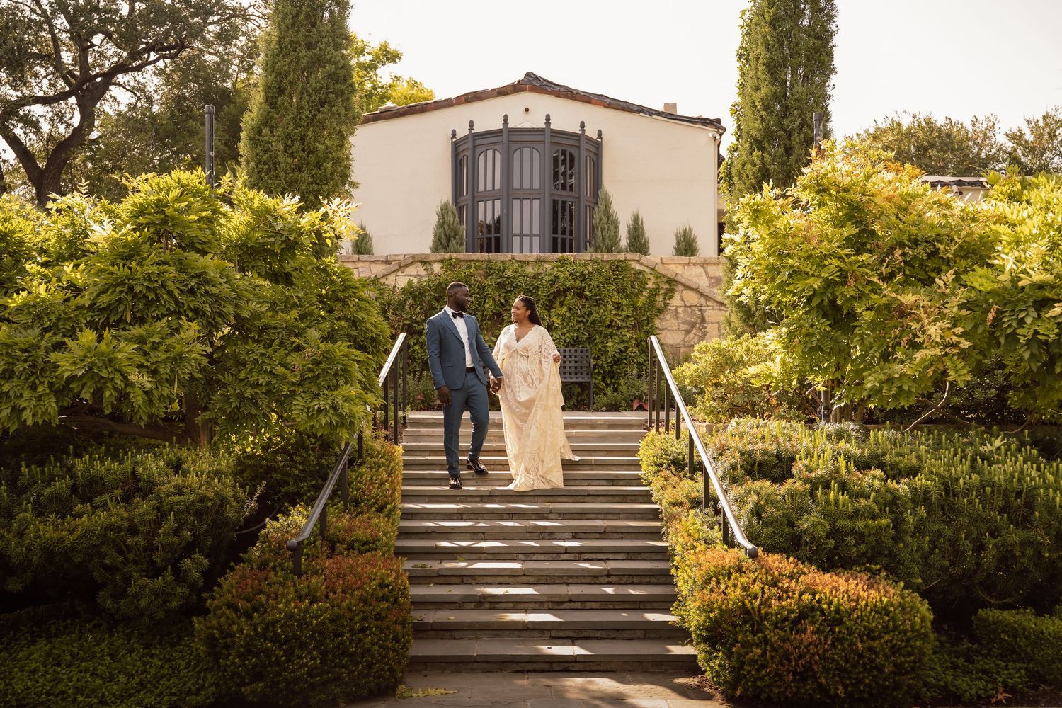 Bride and groom walking down garden steps at elegant outdoor wedding venue with lush greenery and white building backdrop.