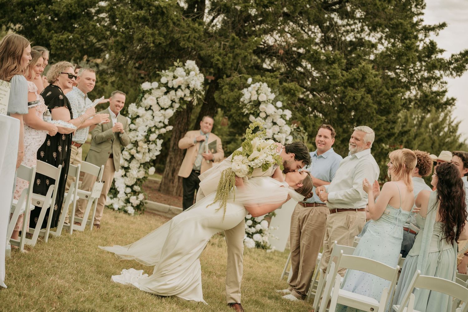 Bride and groom share a romantic dip kiss at outdoor wedding ceremony with floral arch and cheering guests.