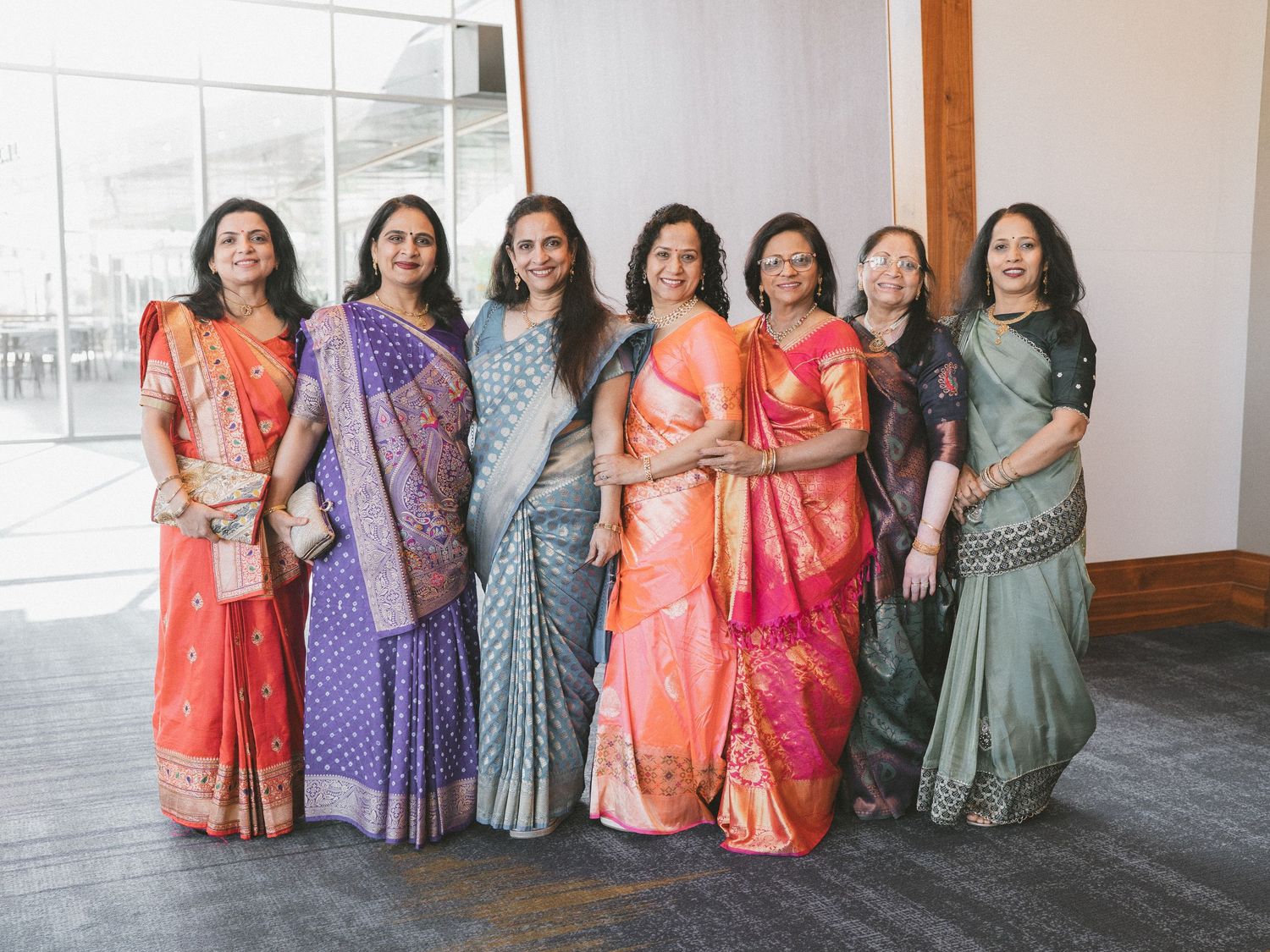 Seven women in vibrant traditional Indian sarees pose together smiling at a formal indoor event.