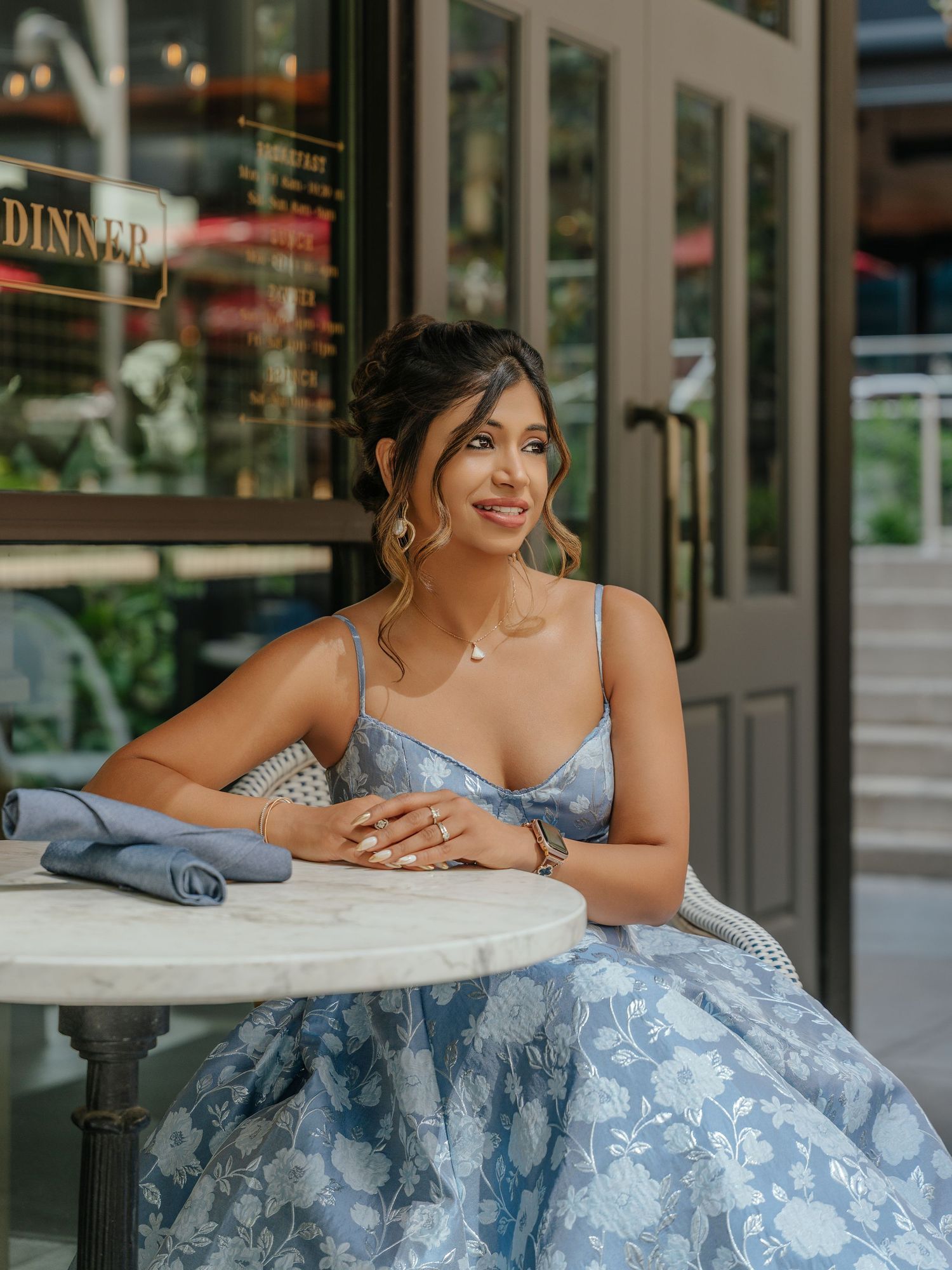 Woman in blue floral dress seated at marble table outside restaurant, smiling elegantly.