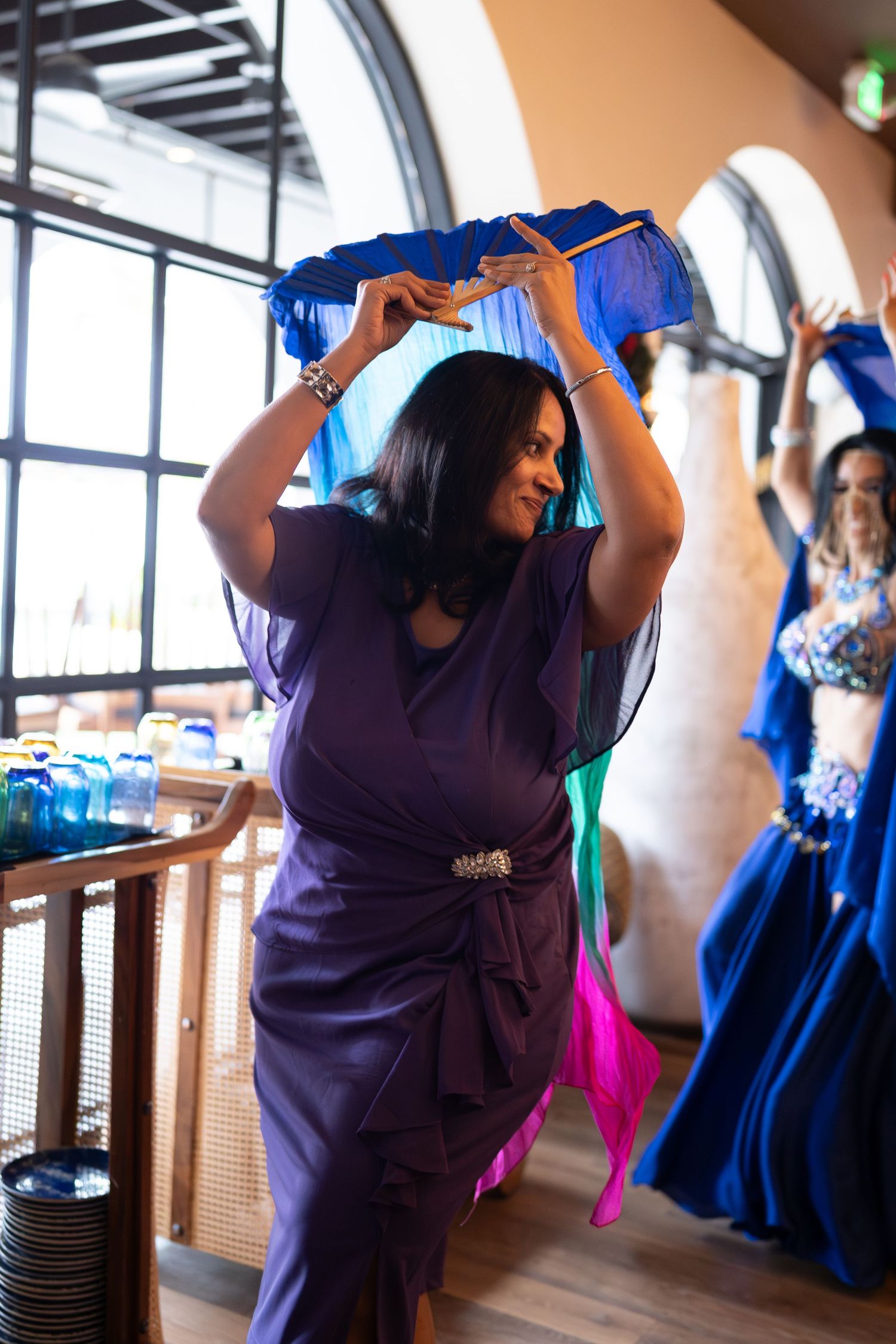 Woman in purple dress holds up blue fabric while dancing at an indoor event with arched windows.