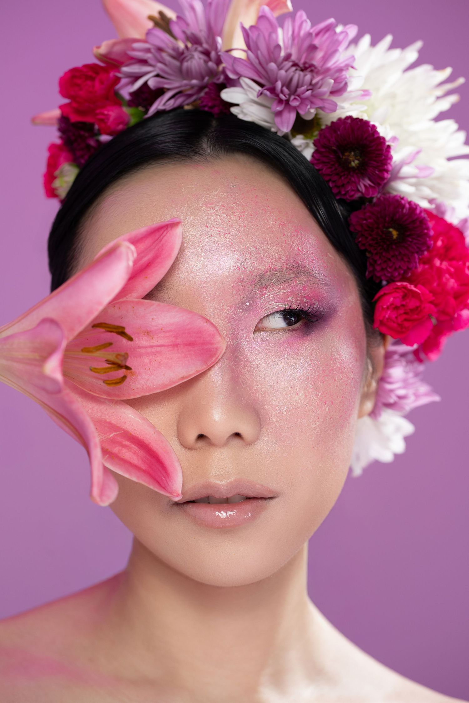 Asian woman with floral crown of pink, purple, and white flowers, holding a pink lily over one eye with glitter makeup.