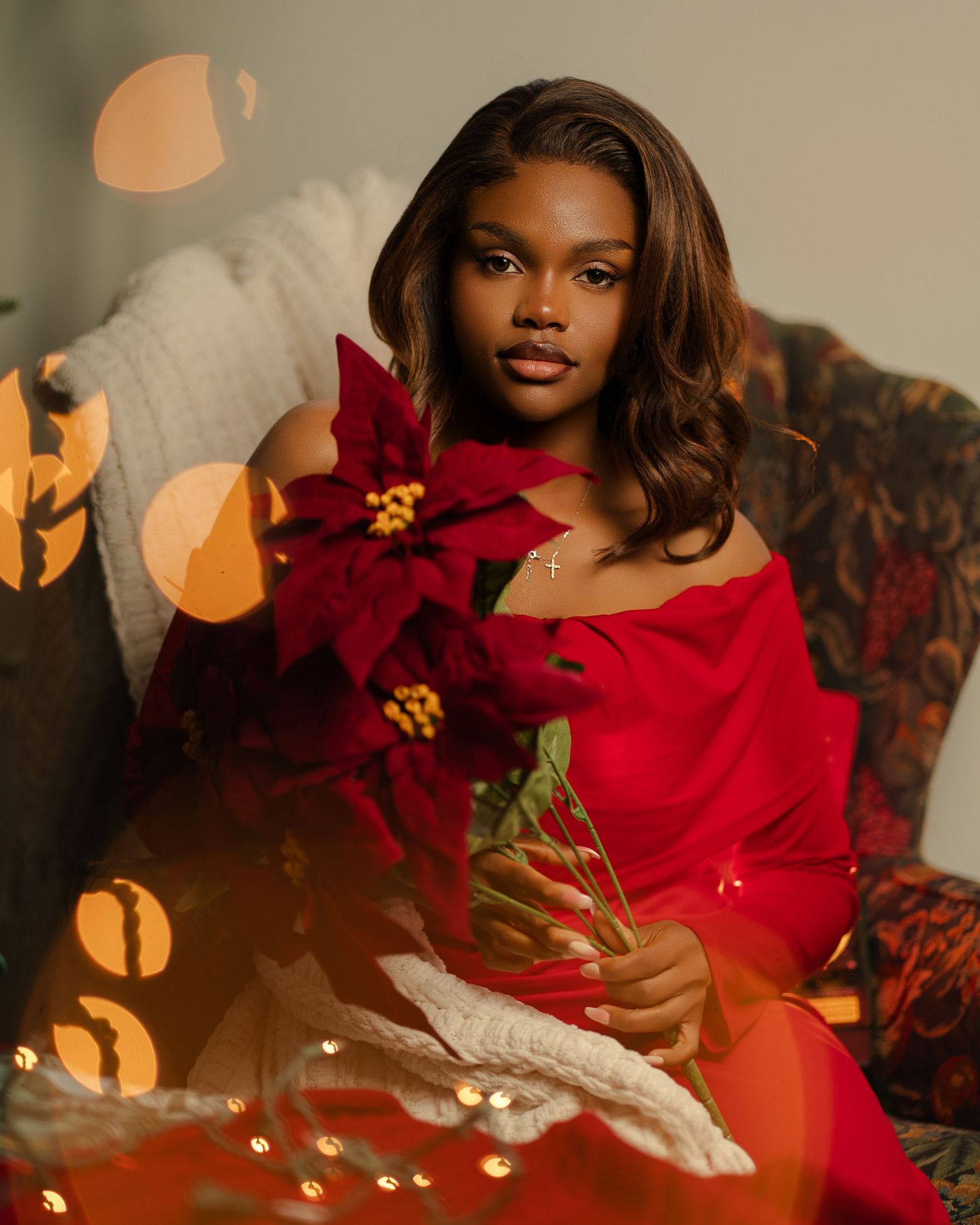 Young woman in red off-shoulder dress holding a red poinsettia flower, surrounded by warm bokeh lights.