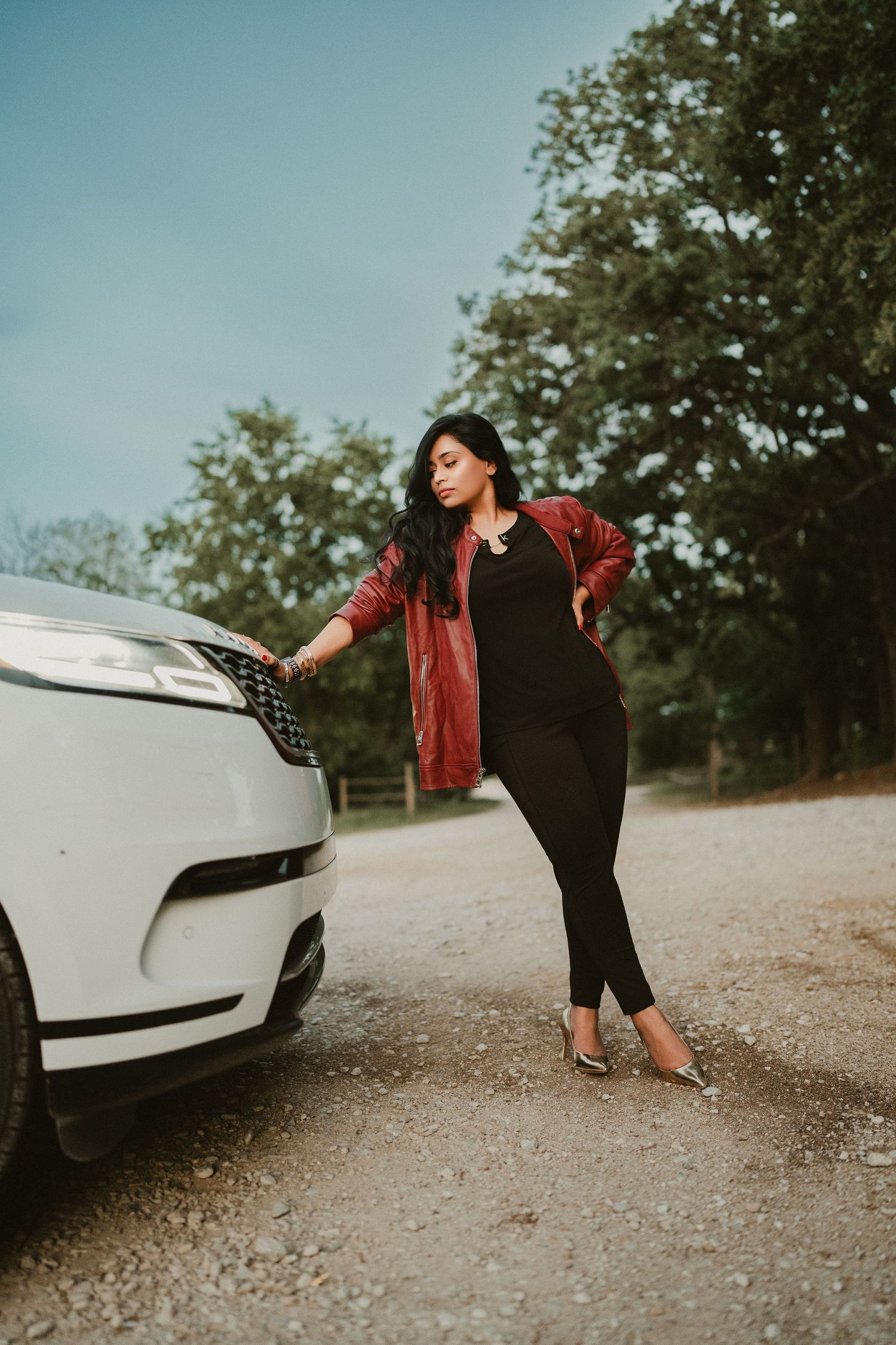 Woman in red jacket and black outfit posing beside a white Range Rover on a gravel road with trees in background.