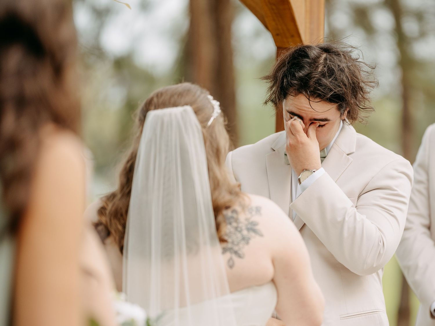 Emotional groom in white suit wiping tears at outdoor wedding ceremony with bride in veil and floral arch.