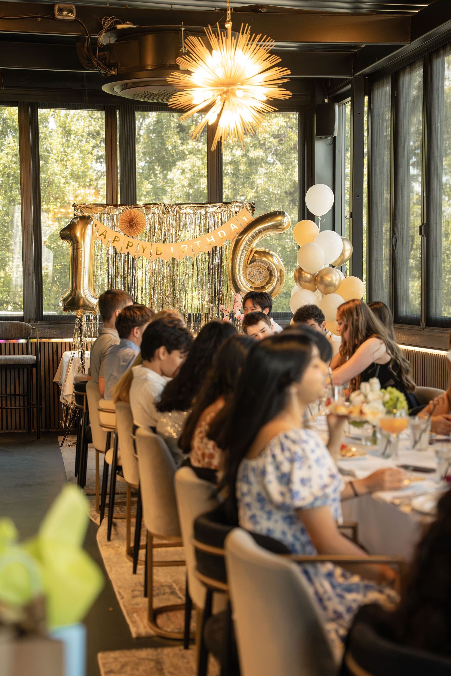 Elegant 16th birthday party with gold balloon numbers, white balloons, and guests seated at decorated tables in a modern venue.