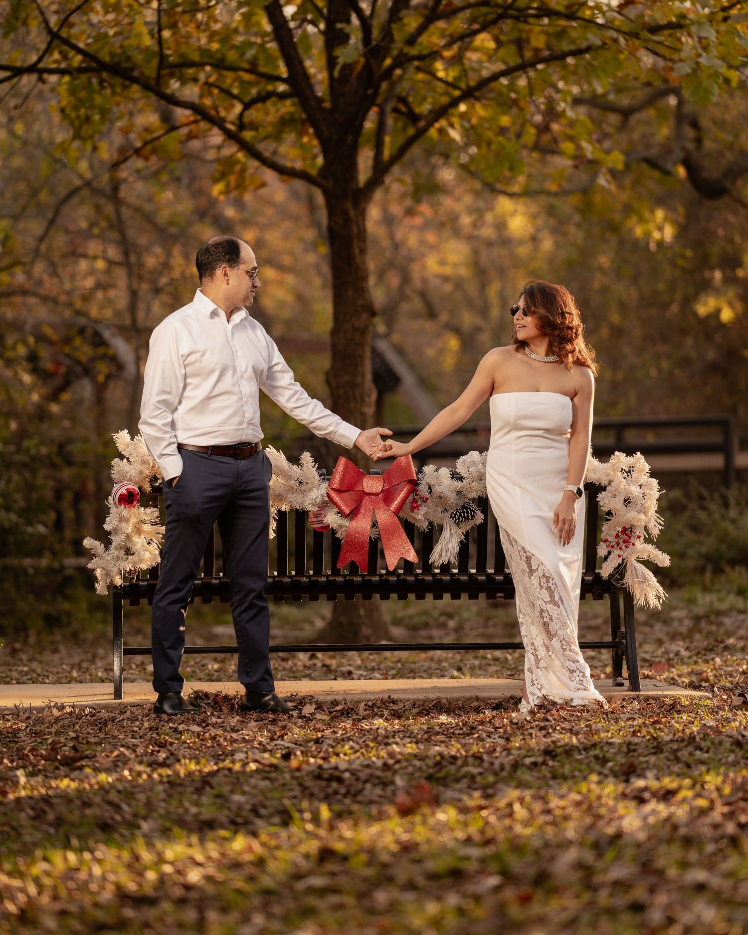 Couple holding hands by a decorated bench with red bow in an autumn park, woman in white lace gown.