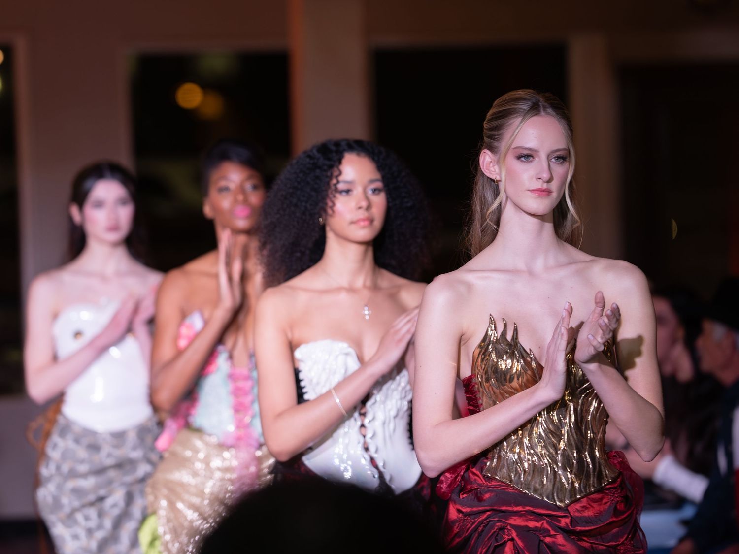 Models walk down a dark runway wearing elegant evening gowns during a high-end fashion show.