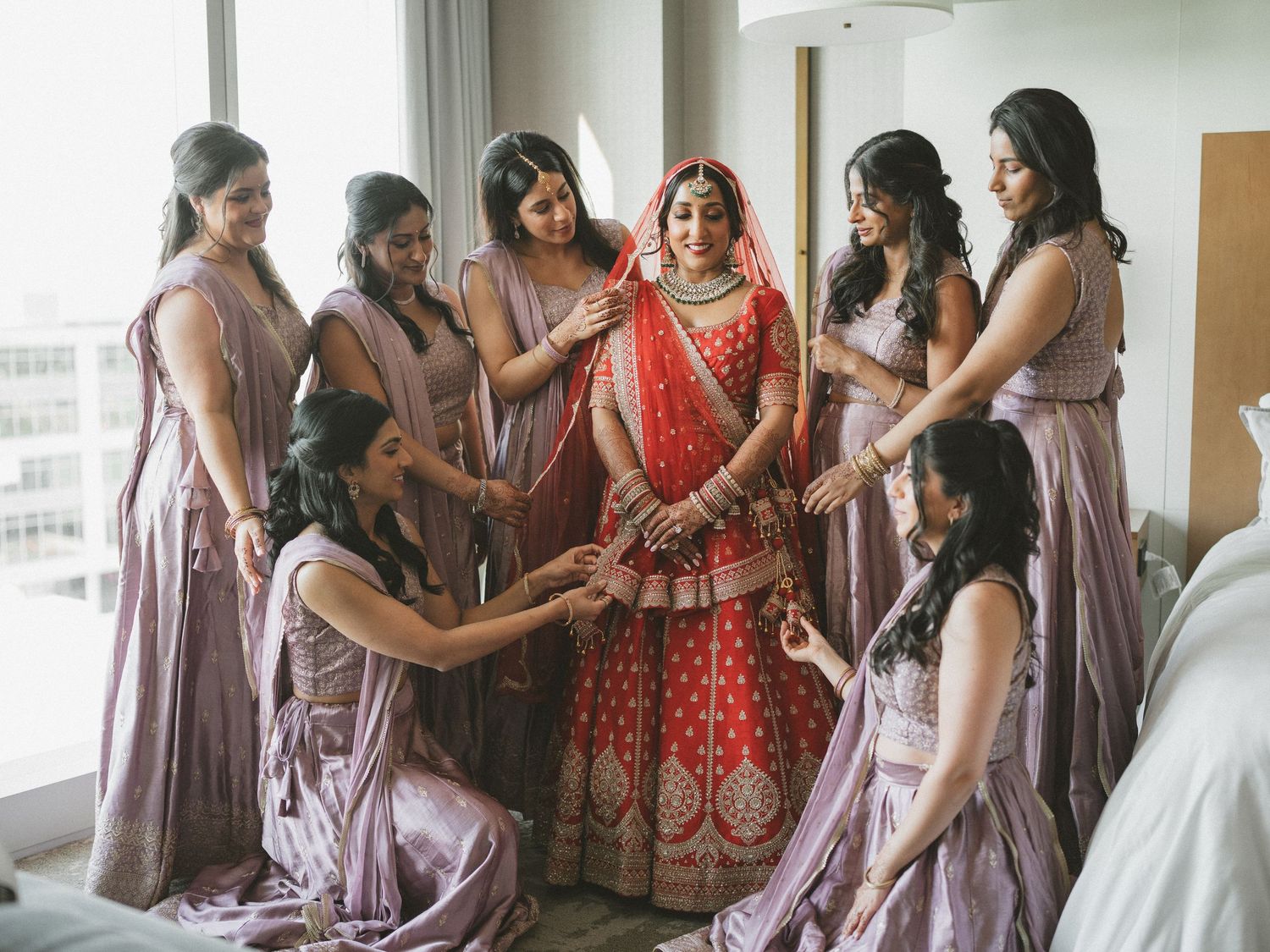 Indian bride in red lehenga surrounded by bridesmaids in lavender sarees during bridal preparation ceremony.